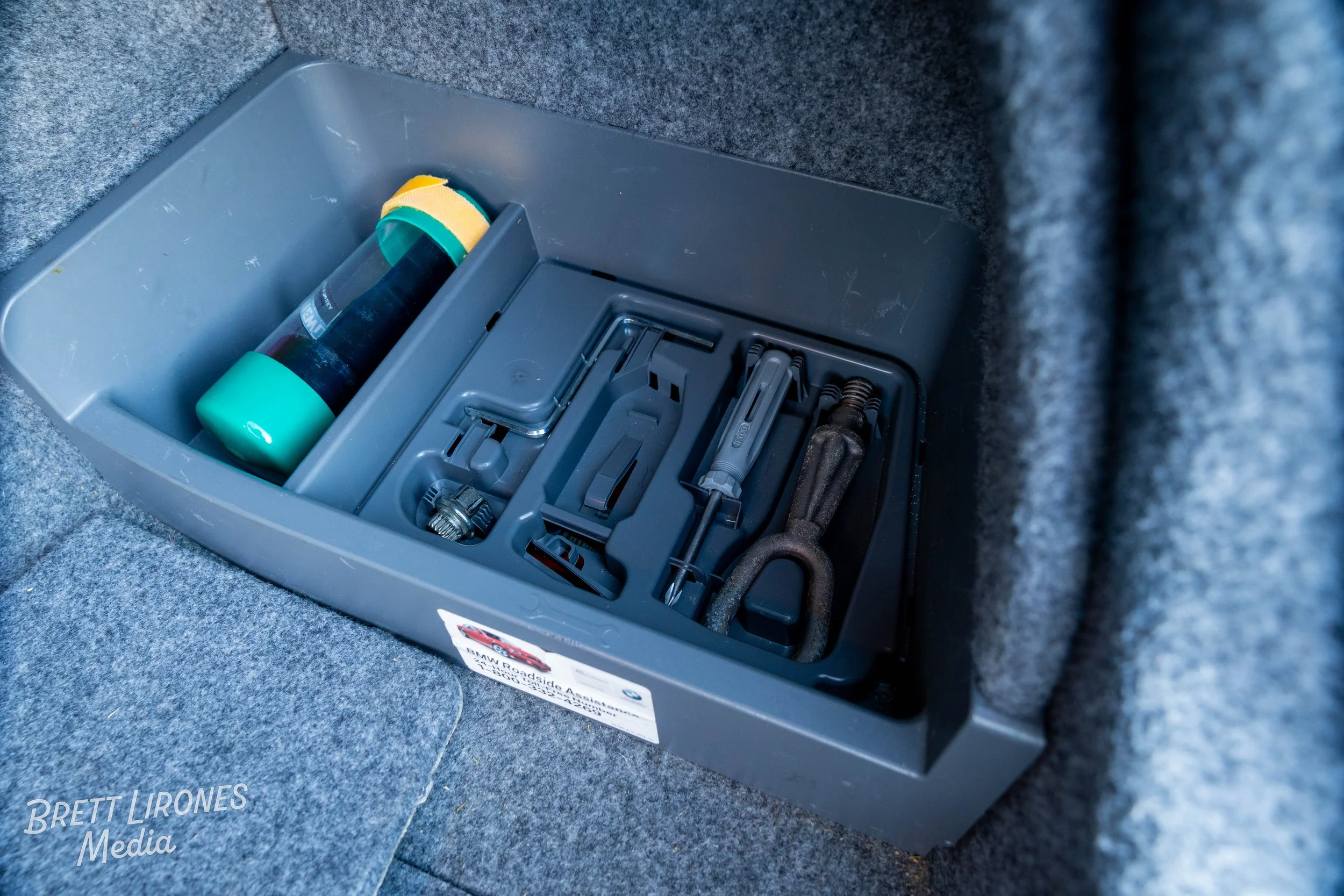 A gray plastic toolbox containing a flashlight, a wrench, screwdrivers, and other tools, placed inside a car trunk on gray carpet.