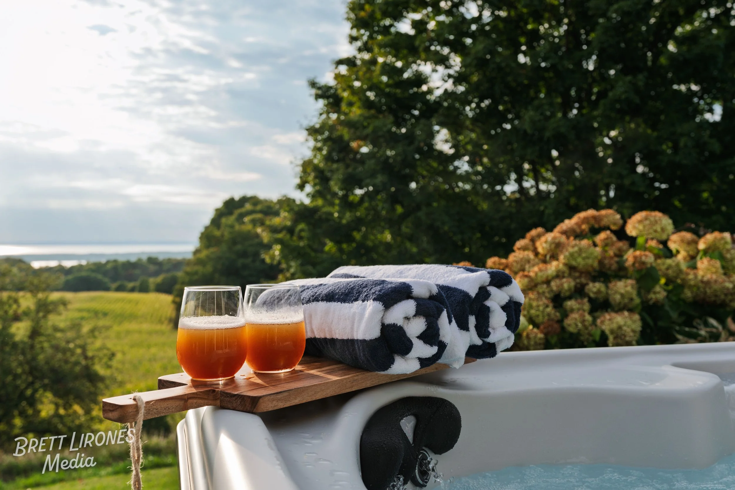 Outdoor hot tub with folded towels, two glasses of orange juice on a wooden tray, greenery, trees, and flowers in the background during daytime.