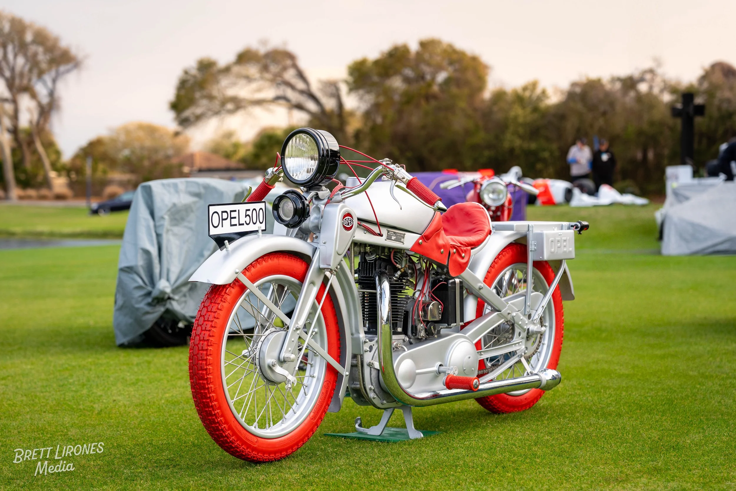 A vintage Opel motorcycle with red and silver body, red tires, and a leather seat, displayed on a grassy field with trees and people in the background.