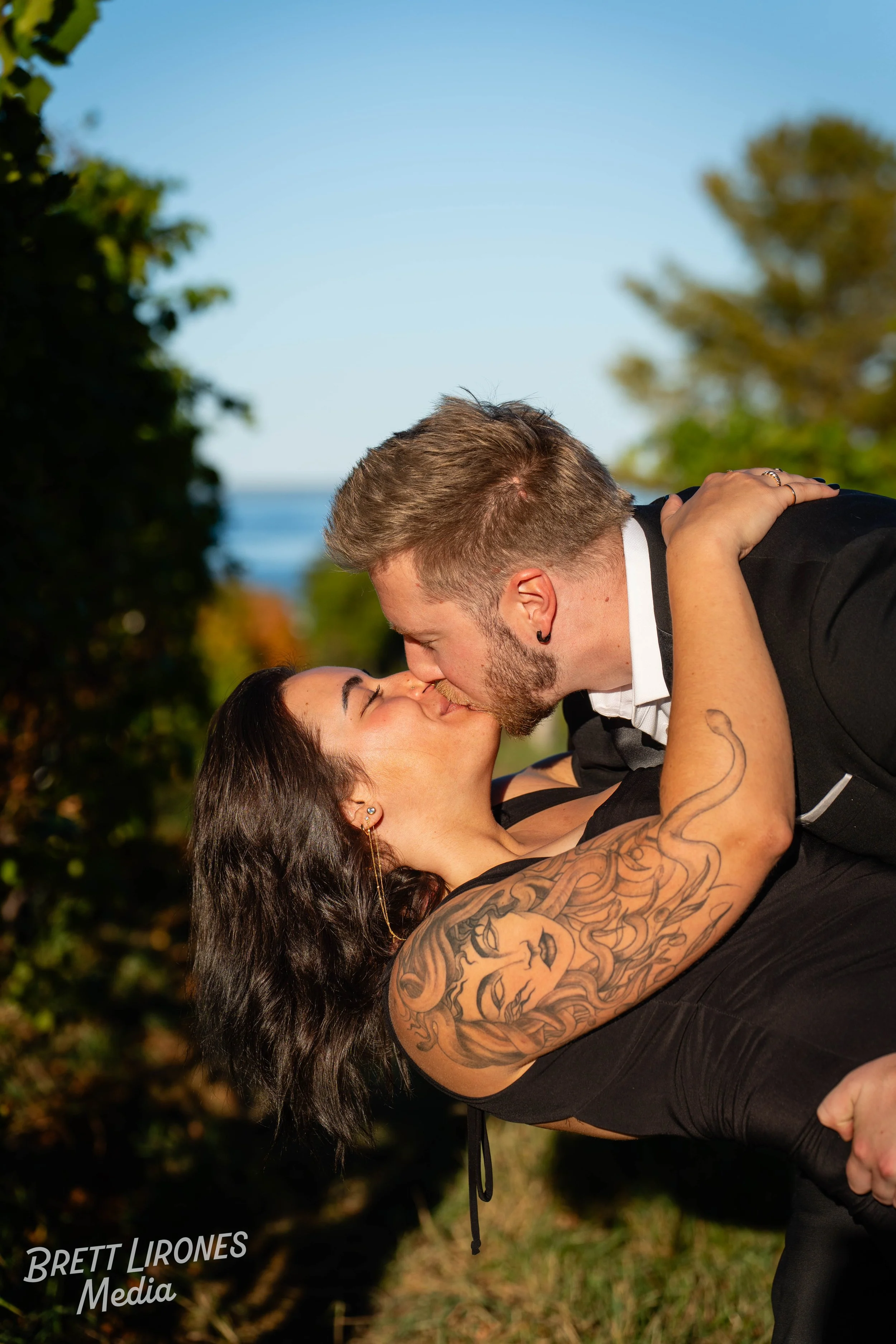 A couple sharing a kiss outdoors during daytime, with the man holding the woman in a dip, both with closed eyes, surrounded by trees and clear blue sky.