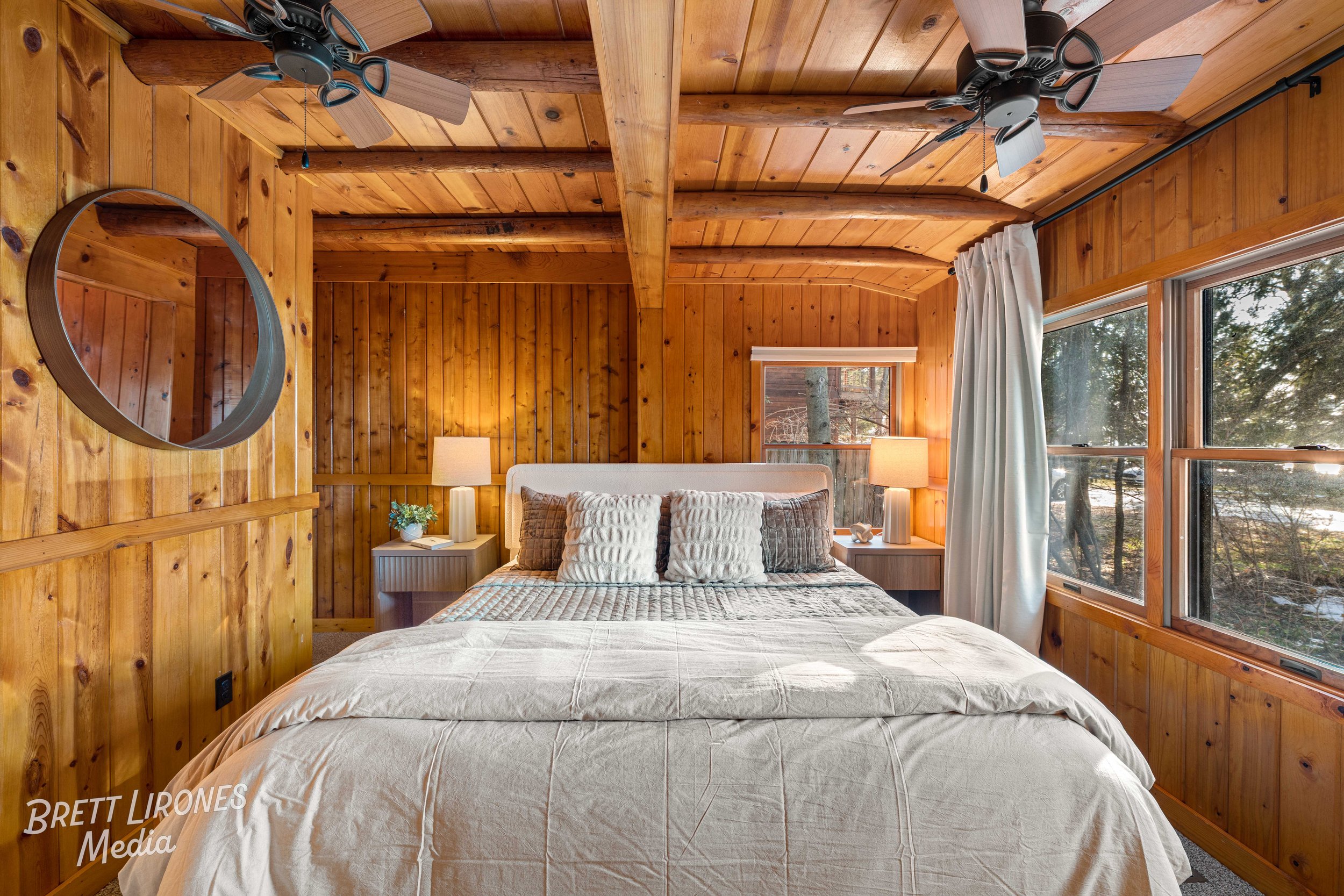 Wood-paneled bedroom with a white bed, throw pillows, bedside lamps, large windows with white curtains, and ceiling fans.