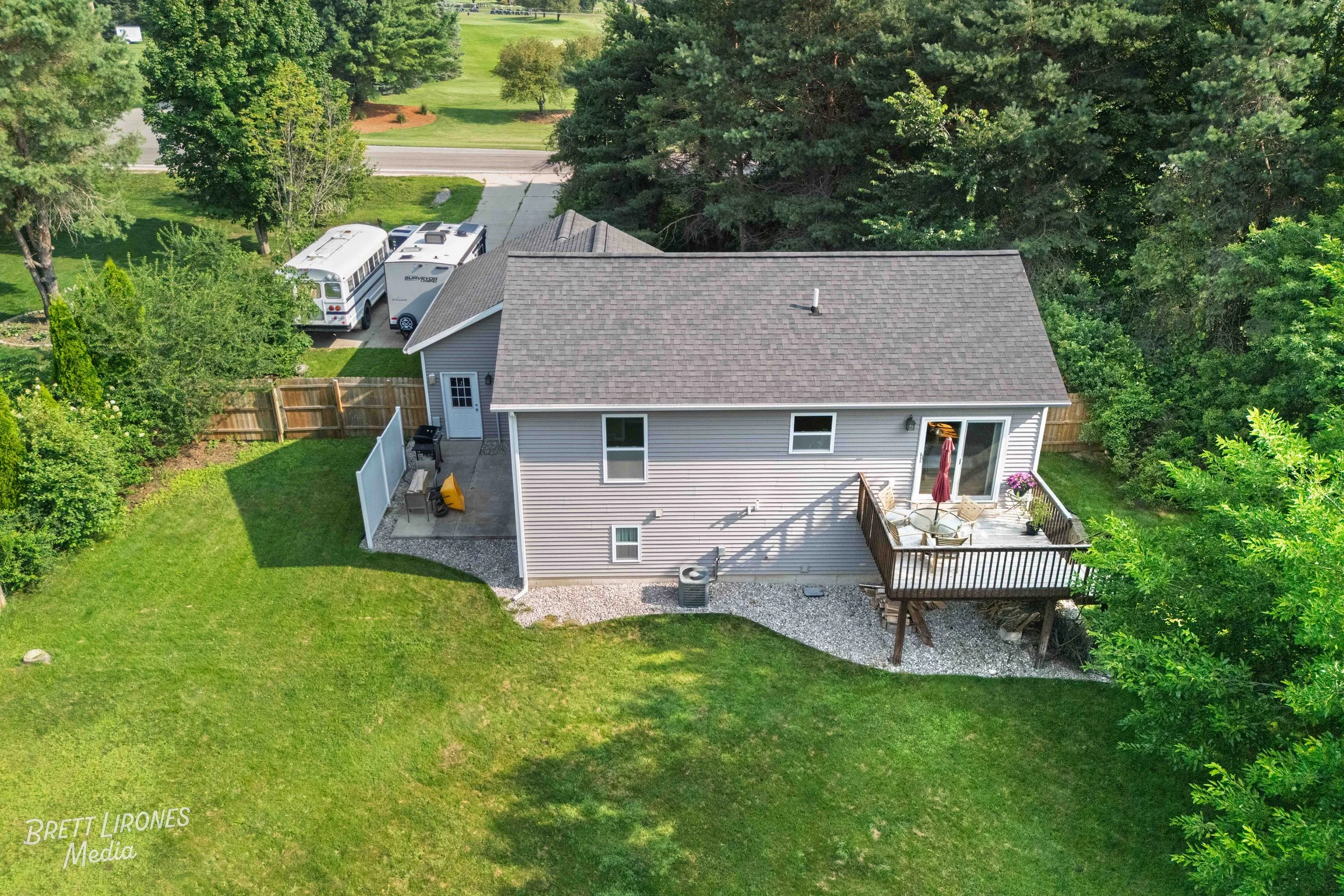 Aerial view of a suburban backyard with a deck, sliding glass door, outdoor furniture, and a small grill. The yard has a grassy area, a gravel pathway, and is bordered by wooden fences and surrounded by trees.