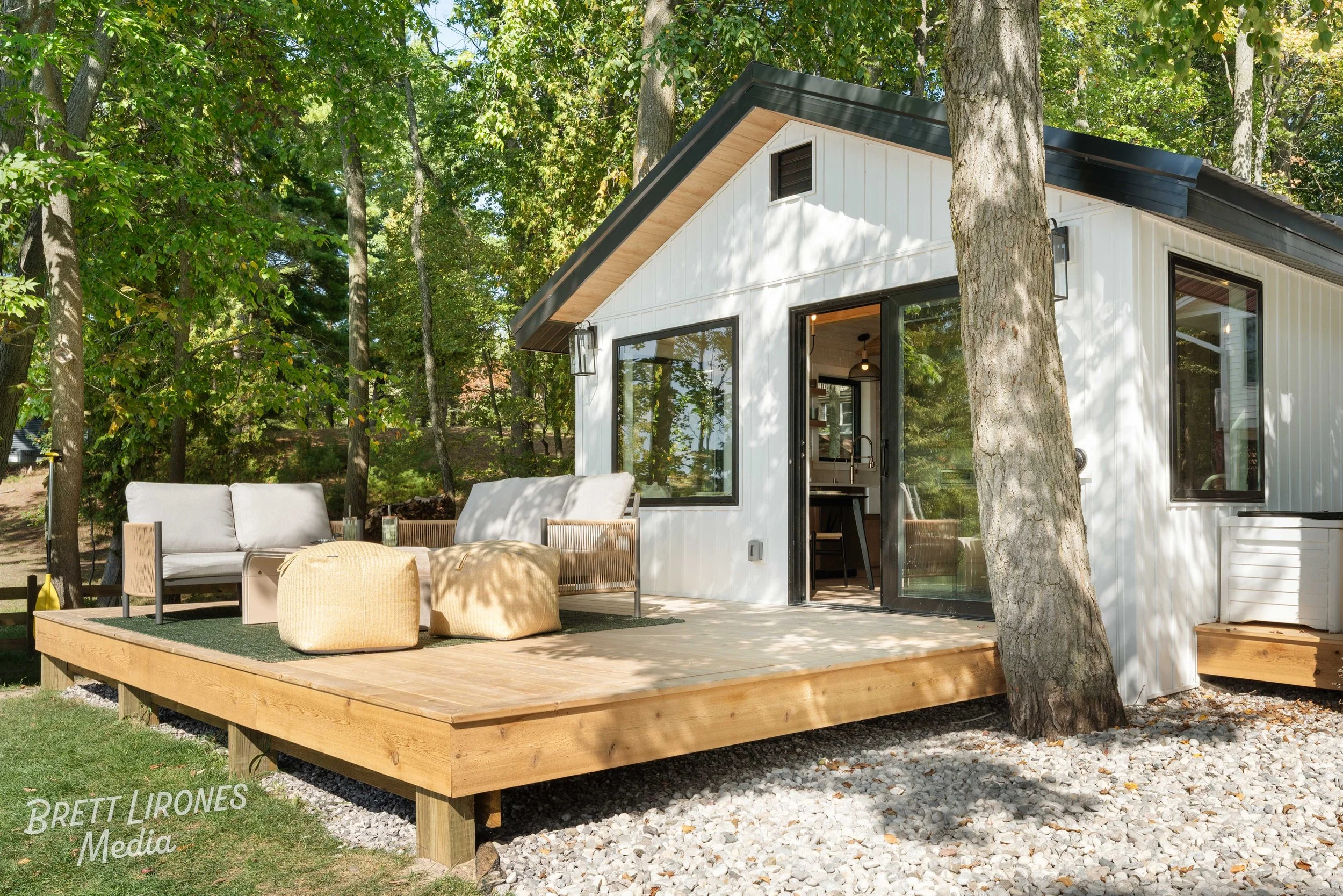 Modern white house with large glass windows and sliding doors, surrounded by trees, featuring a wooden deck with outdoor seating and cushions.