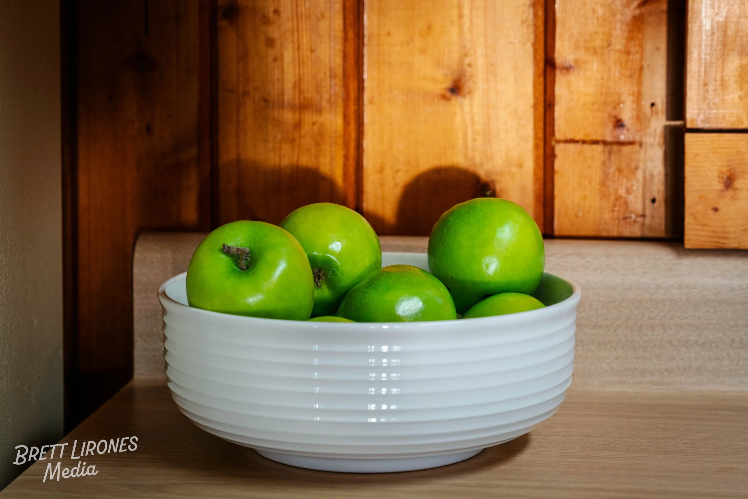 A white bowl filled with bright green apples on a wooden surface with a wood-paneled wall in the background.