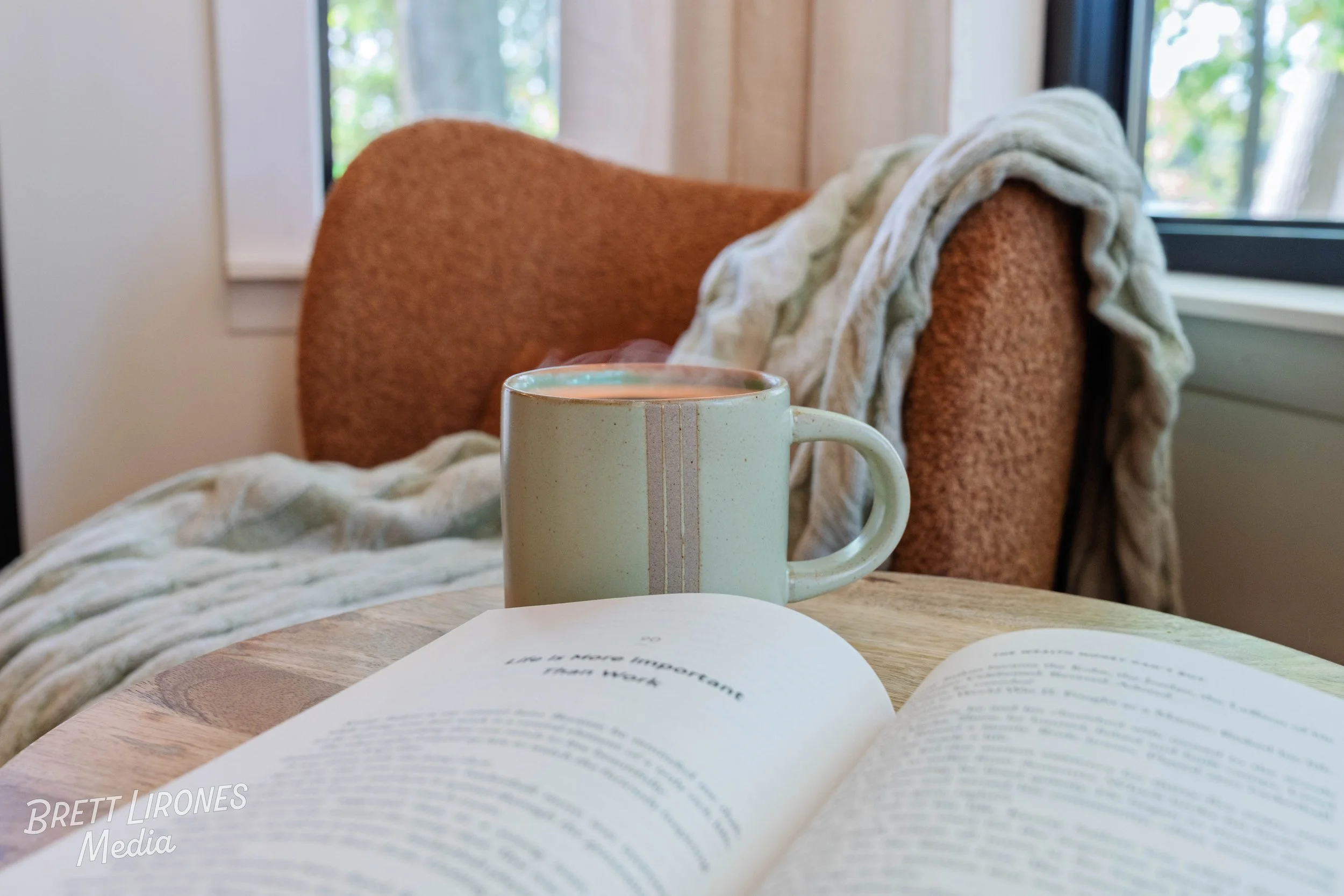 A steaming cup of coffee or tea on a wooden table next to an open book with blurry text, a cushioned chair with a blanket, and a window with trees outside.