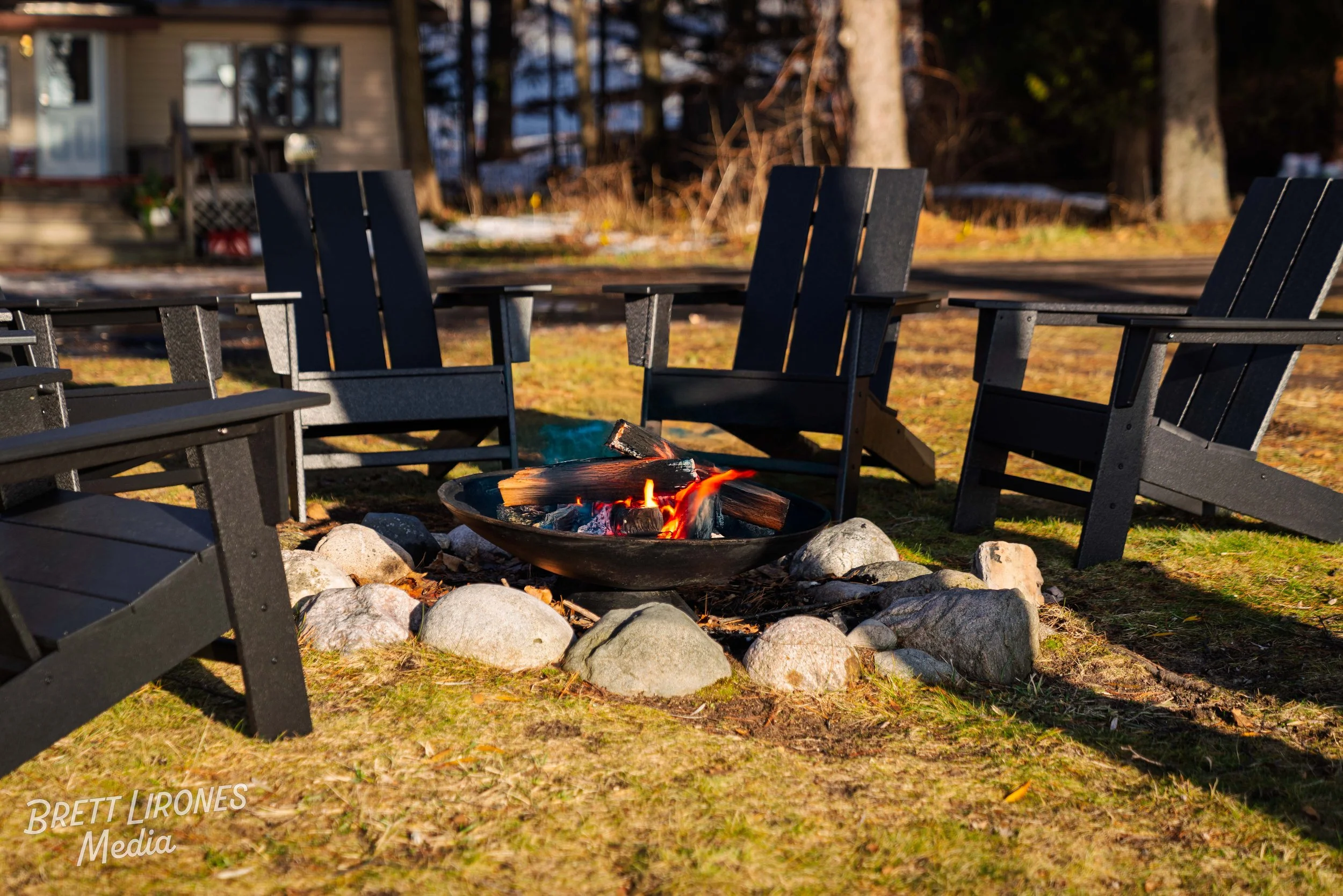 Four black outdoor chairs arranged around a fire pit with burning logs, set on a grassy area with rocks surrounding the fire, and a house and trees in the background.
