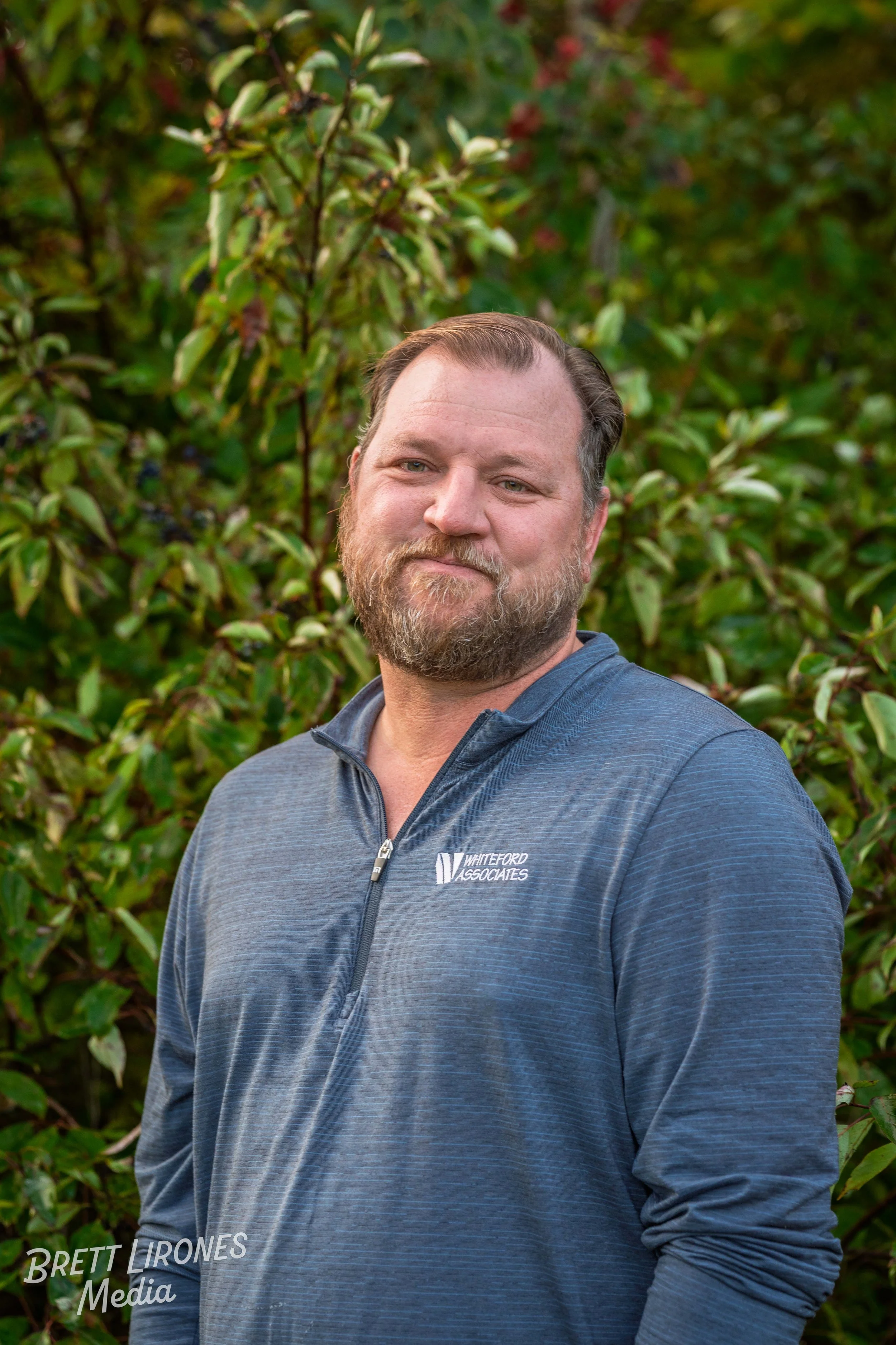 A bearded man with light skin standing outdoors in front of green foliage, wearing a blue quarter-zip shirt with the logo 'Whiteford Associates'.