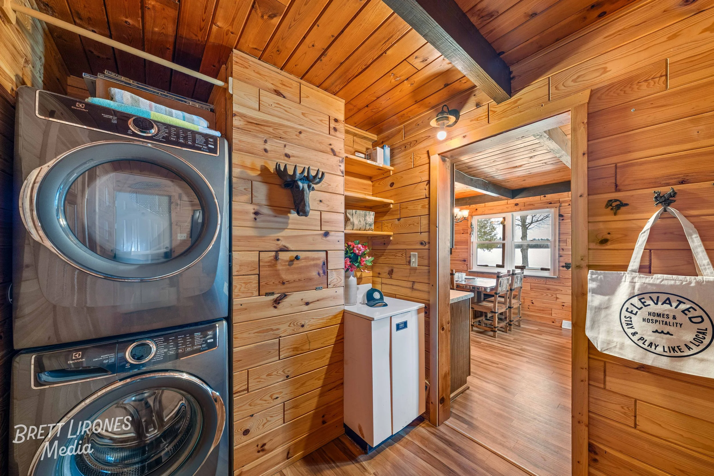 Interior of a rustic wooden cabin laundry area with a stacked washer and dryer on the left, a wall-mounted decorative moose head, and a doorway leading to a dining space with windows and trees visible outside.