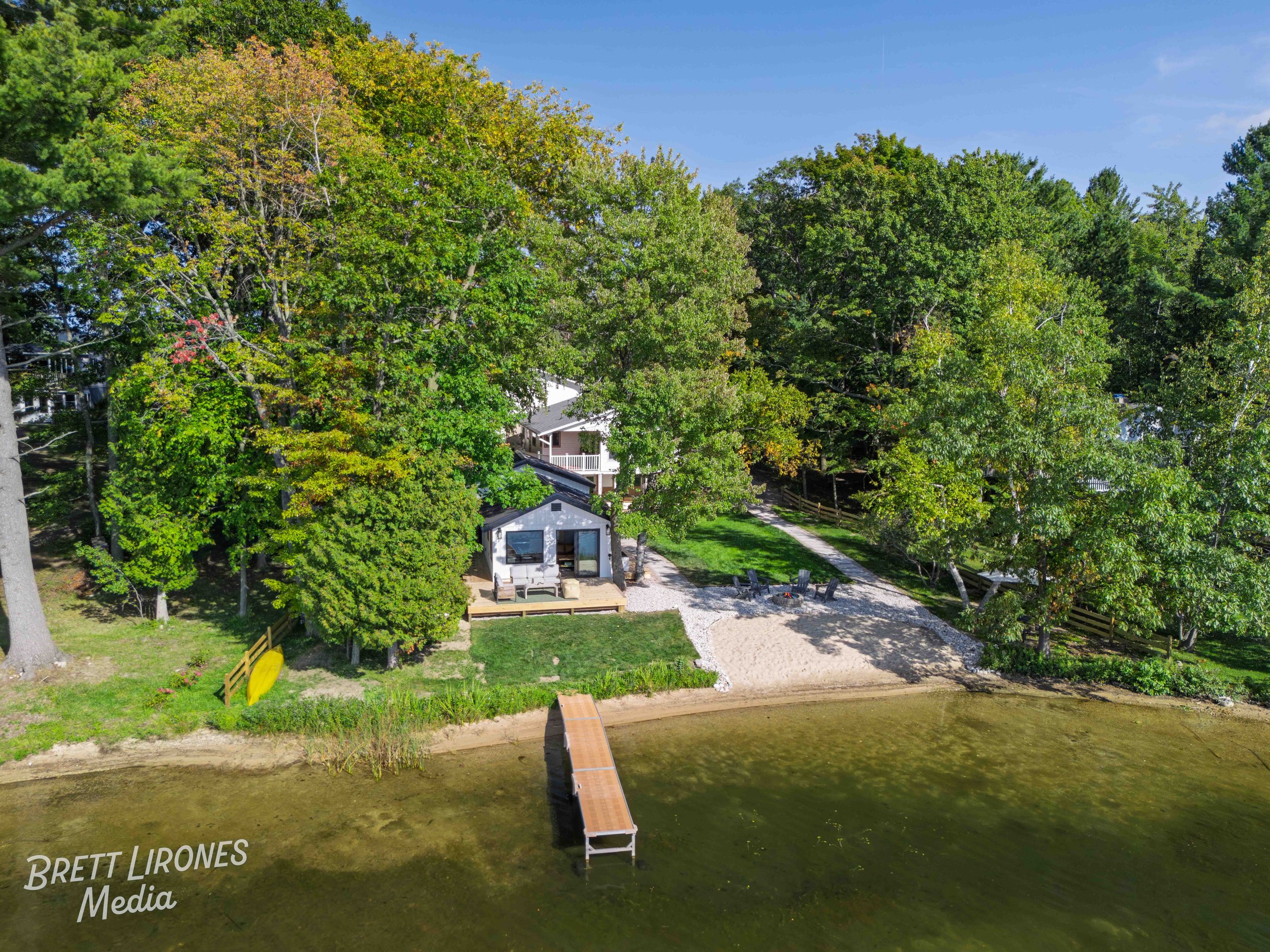 Aerial view of a lakeside property with trees, a small house, a gravel area with outdoor furniture, and a dock extending into the water.