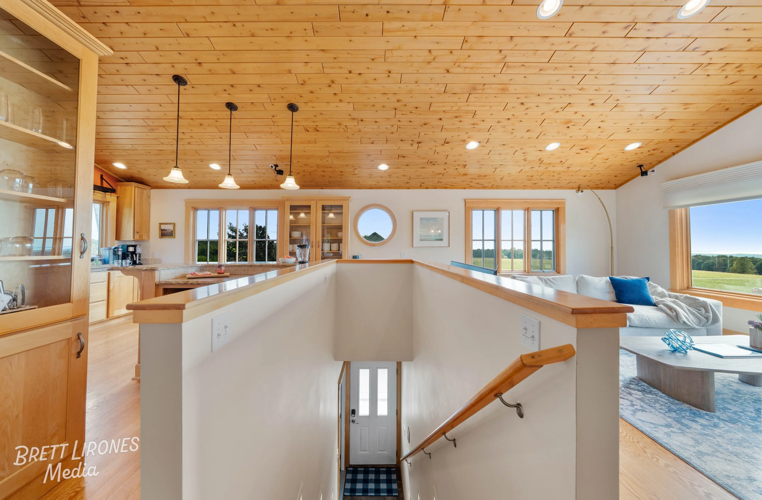 Interior view of a house with a wooden ceiling, open kitchen with granite countertops, large windows showing a green landscape, and a staircase leading downstairs.