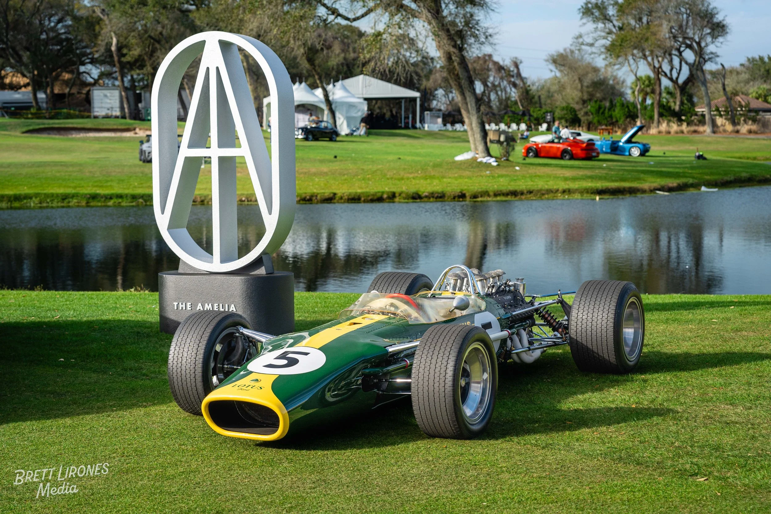 A vintage green and yellow race car with the number 5 on the front, parked on grass near a body of water at an outdoor event. A large modern sculpture with an MMA logo and the words 'The Amelia' is in the background, along with trees, tents, and othe