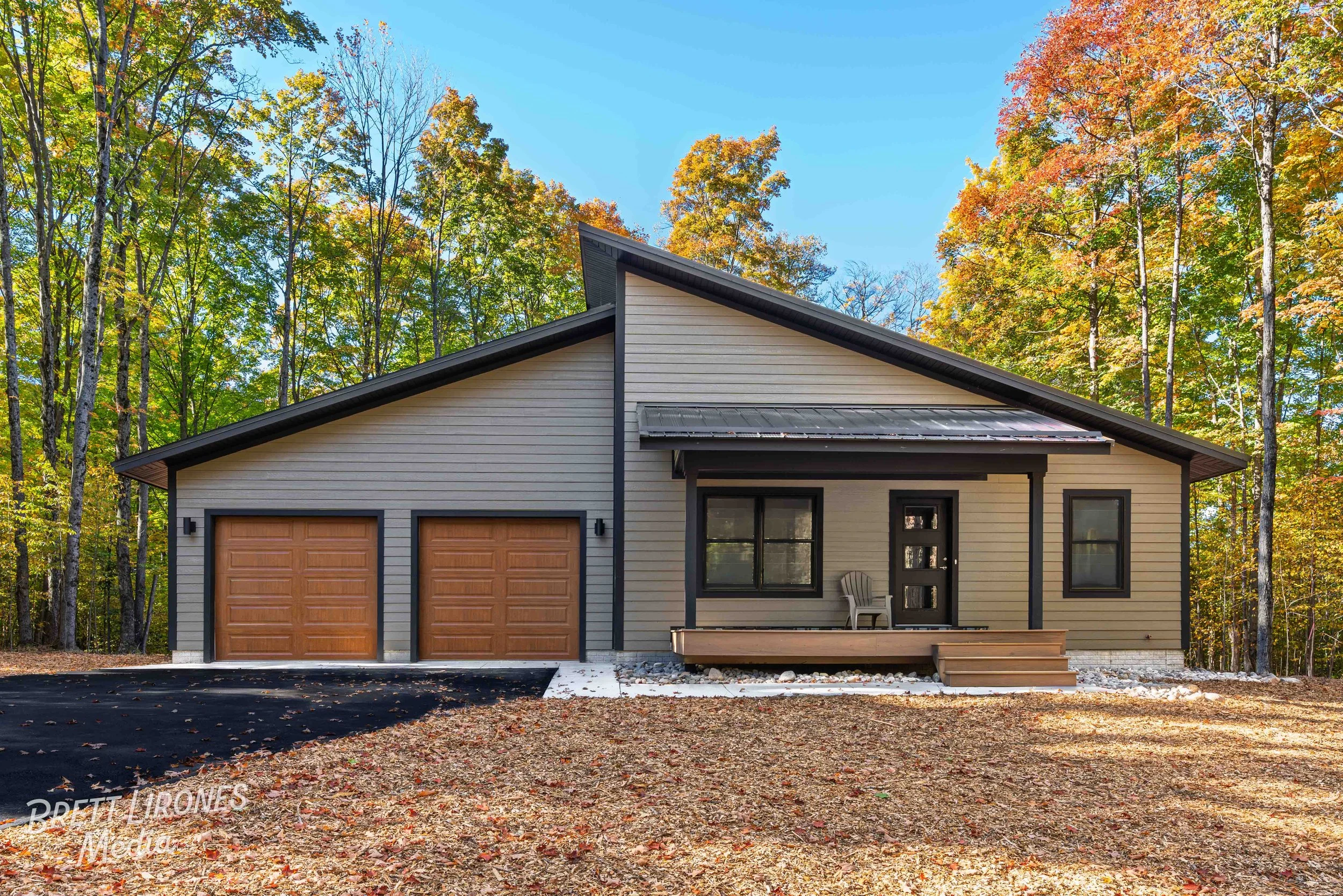 Modern house with beige siding, two wooden garage doors, black trim, and a small front porch with steps, surrounded by autumn trees with colorful foliage.