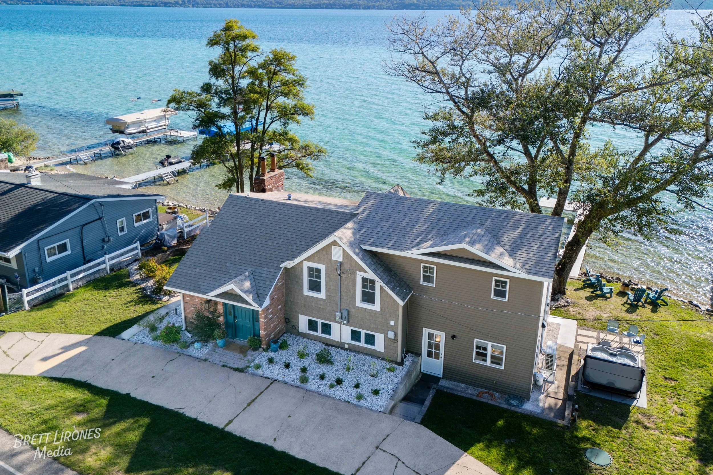 Aerial view of a house next to a lake with docks, boats, and outdoor seating, surrounded by trees and green grass.