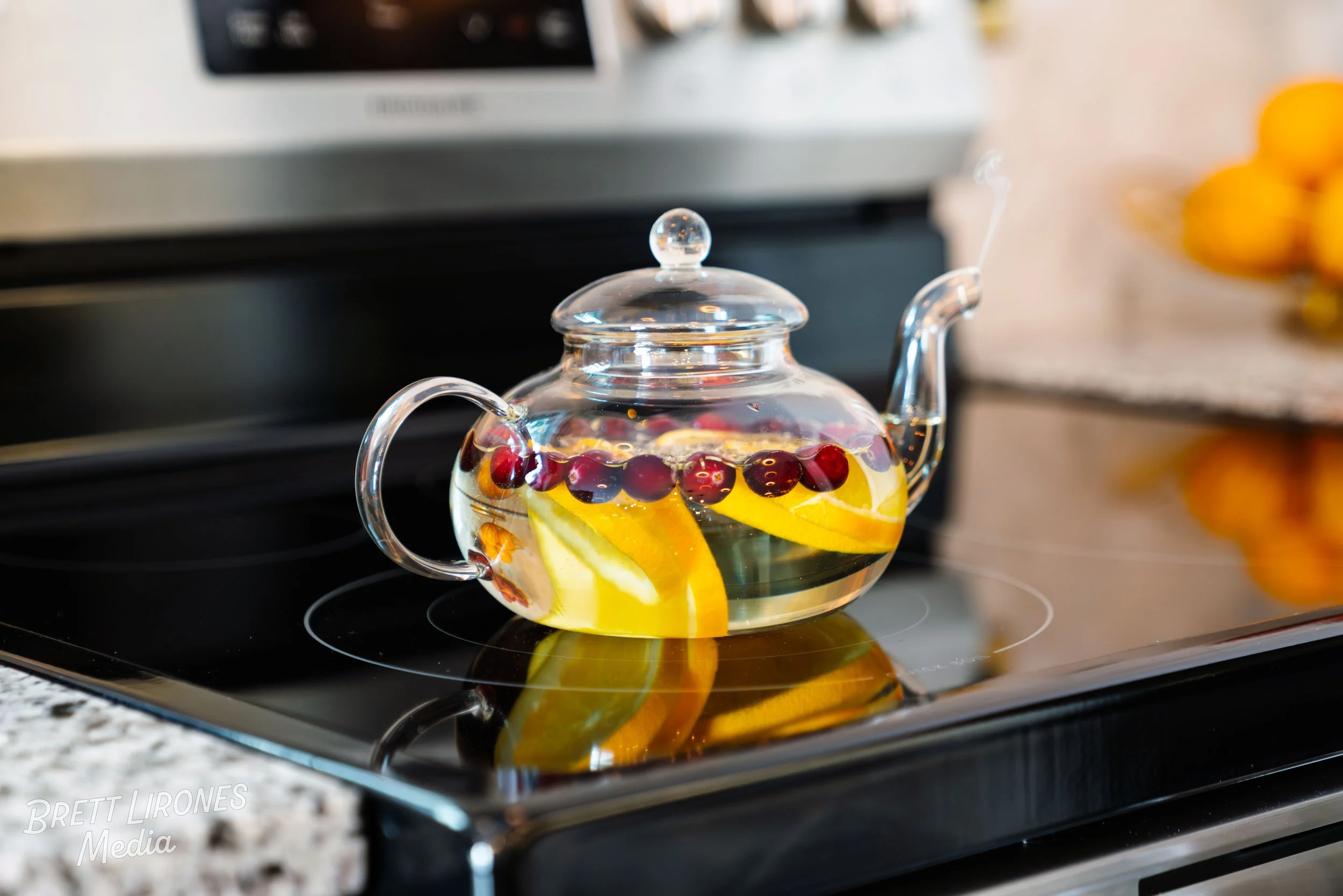 A glass teapot filled with water, lemon slices, and cranberries on a stovetop.