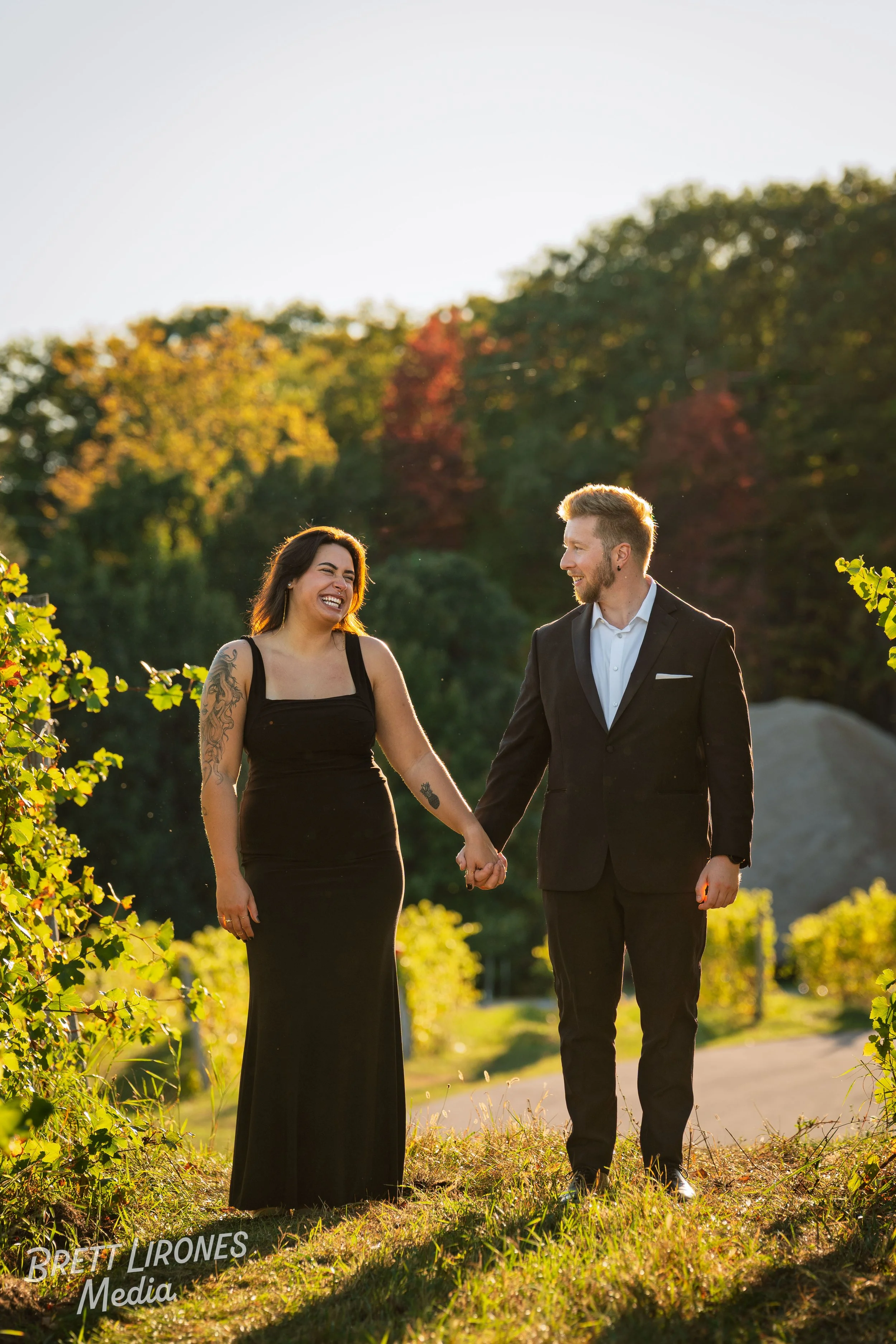 A couple in formal attire holding hands and smiling in a scenic outdoor setting with trees and sunlight.