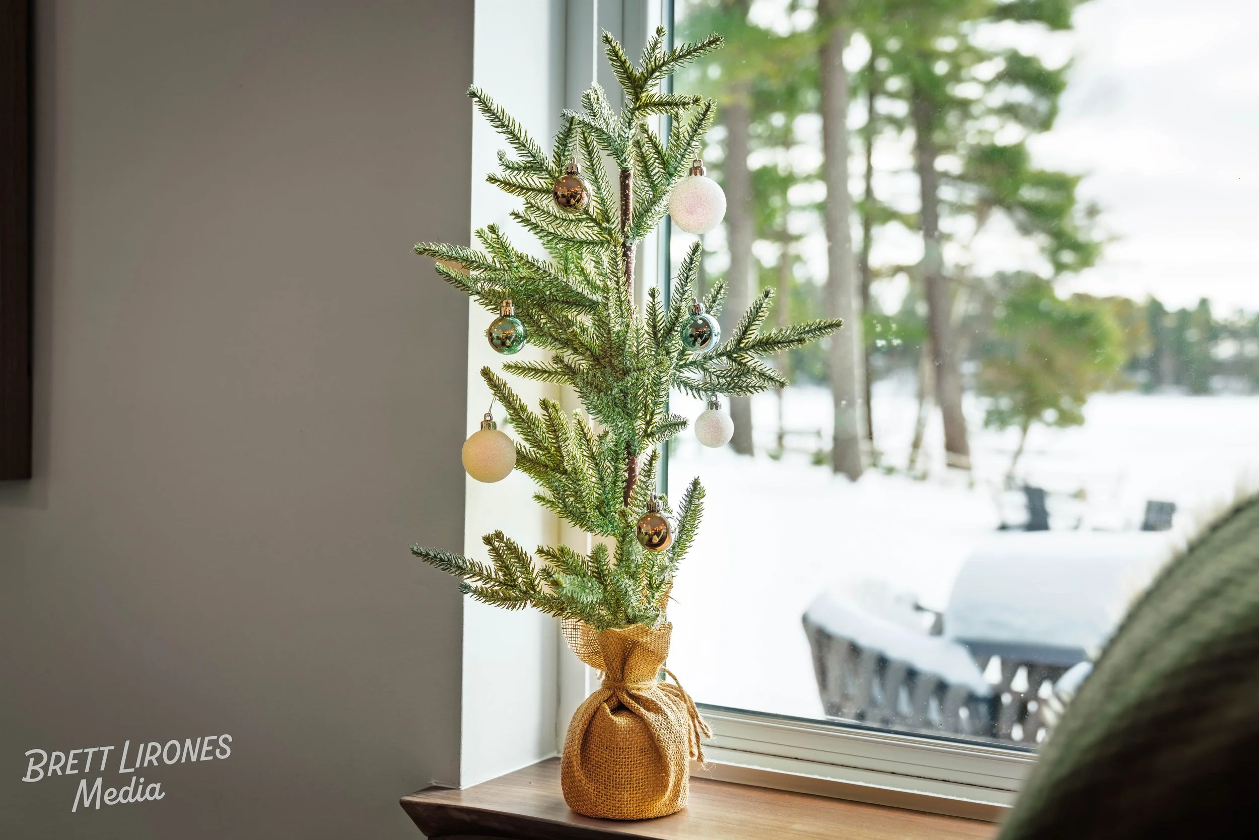 Small Christmas tree decorated with white, gold, and silver ornaments placed on a windowsill, with a snowy outdoor scene visible through the window.