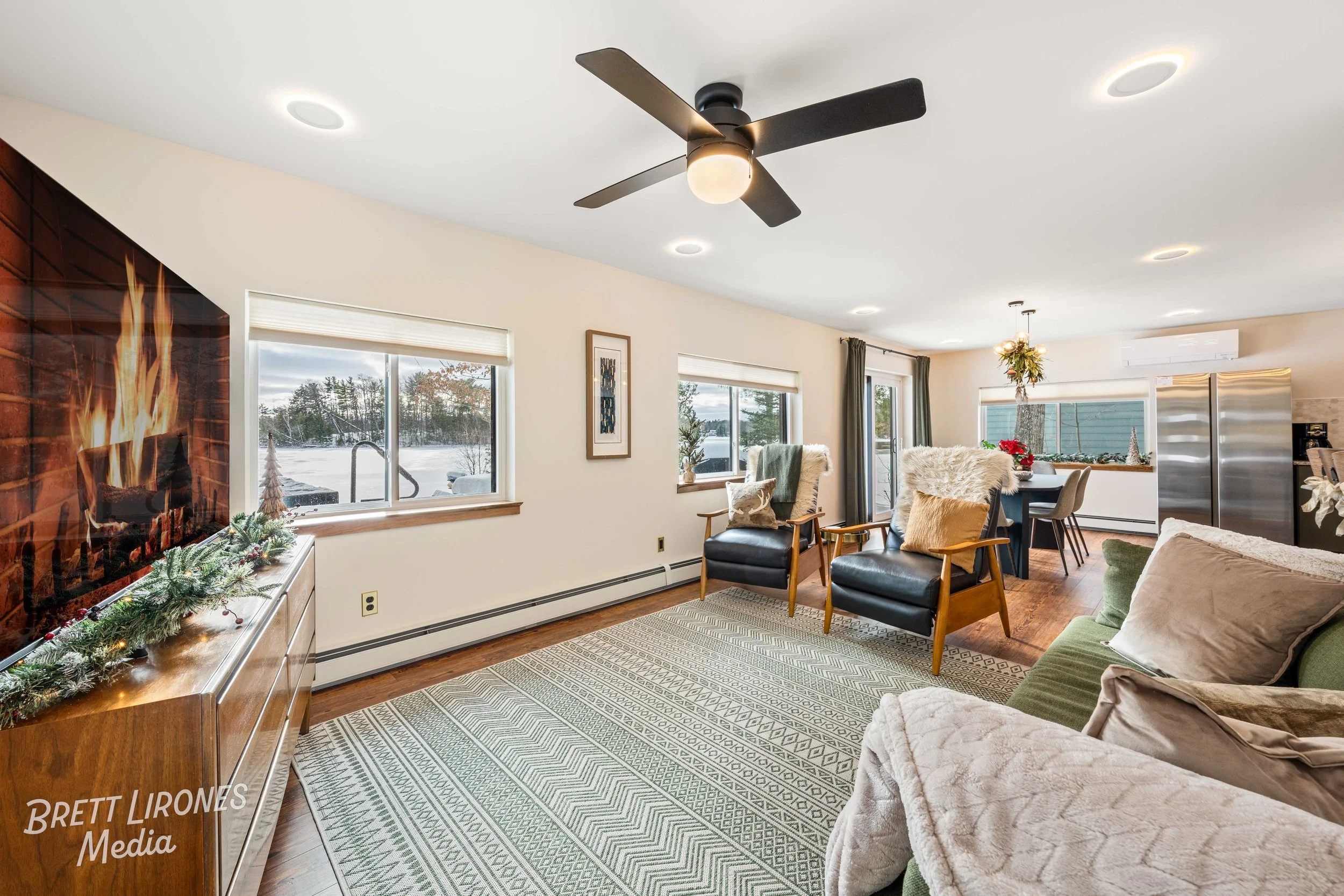 Living room decorated for Christmas with a fireplace, a TV, yellow and green chairs, a glass dining table with chairs, a refrigerator, and windows showing a snowy outdoor landscape.