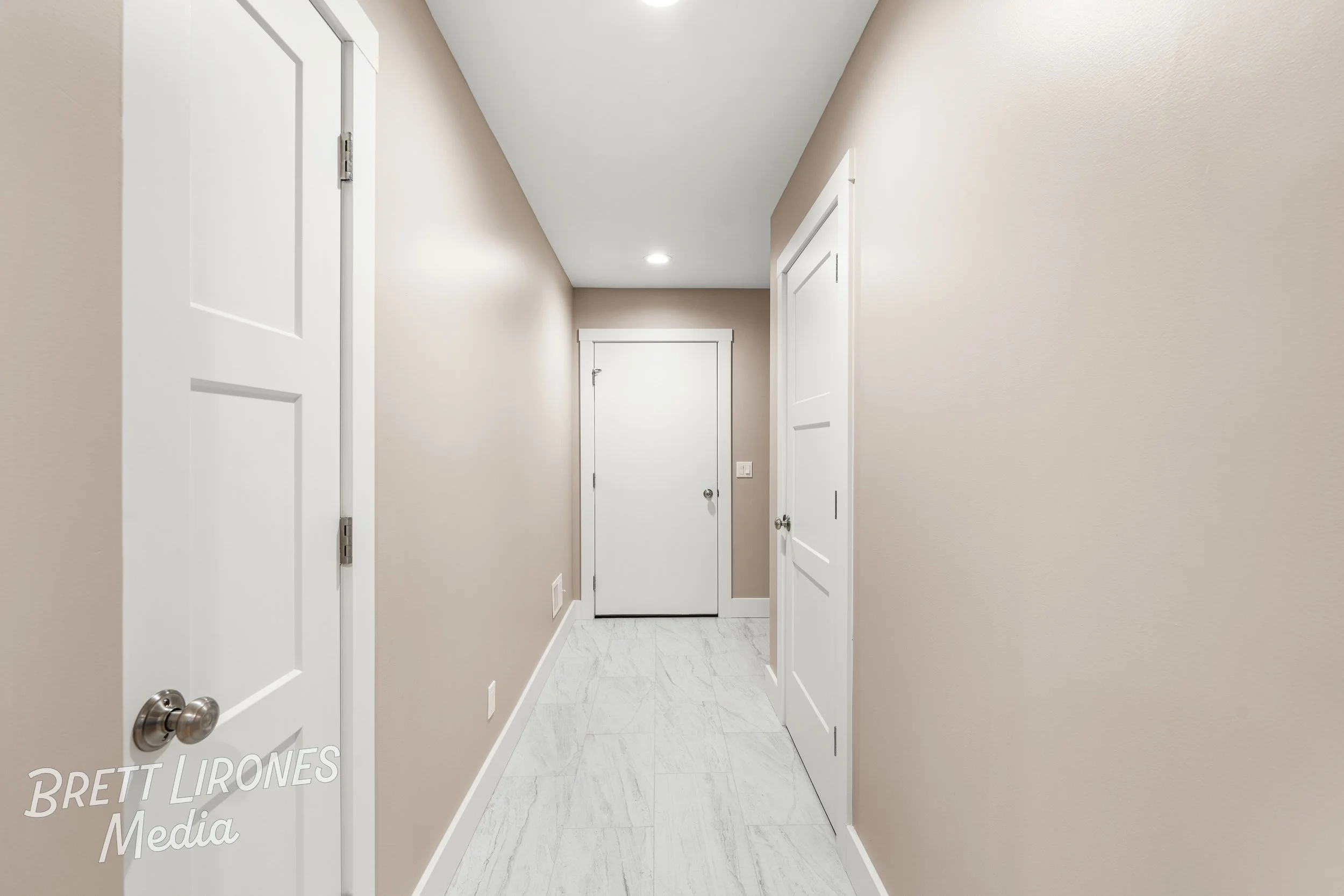 Empty hallway with beige walls, white doors, white baseboards, and white tile flooring leading to a closed door at the end.