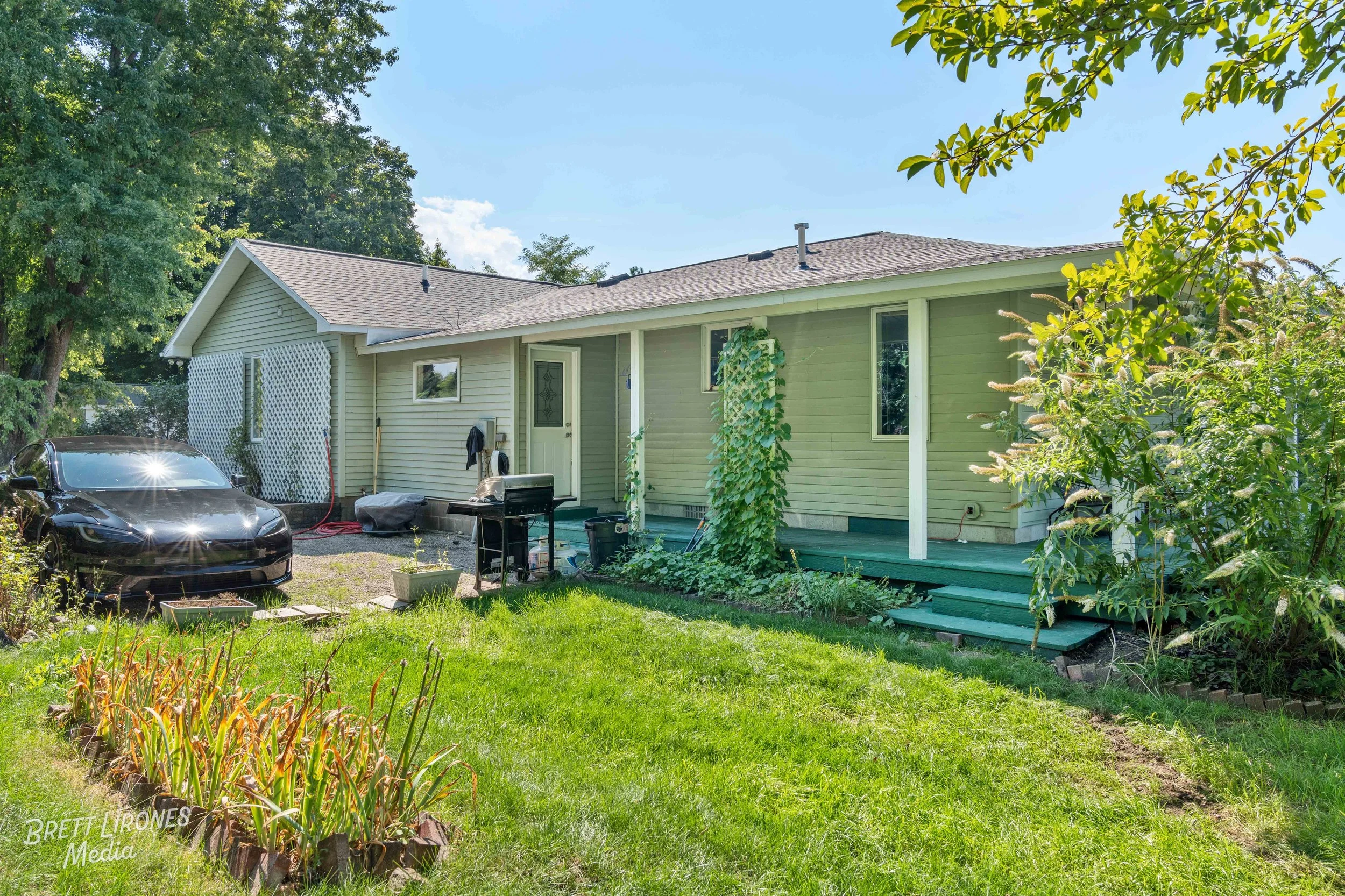 Backyard of a house with a green lawn, a black car parked near the house, and garden plants and vines climbing up the house wall. There are steps leading to a back door and trees in the background.