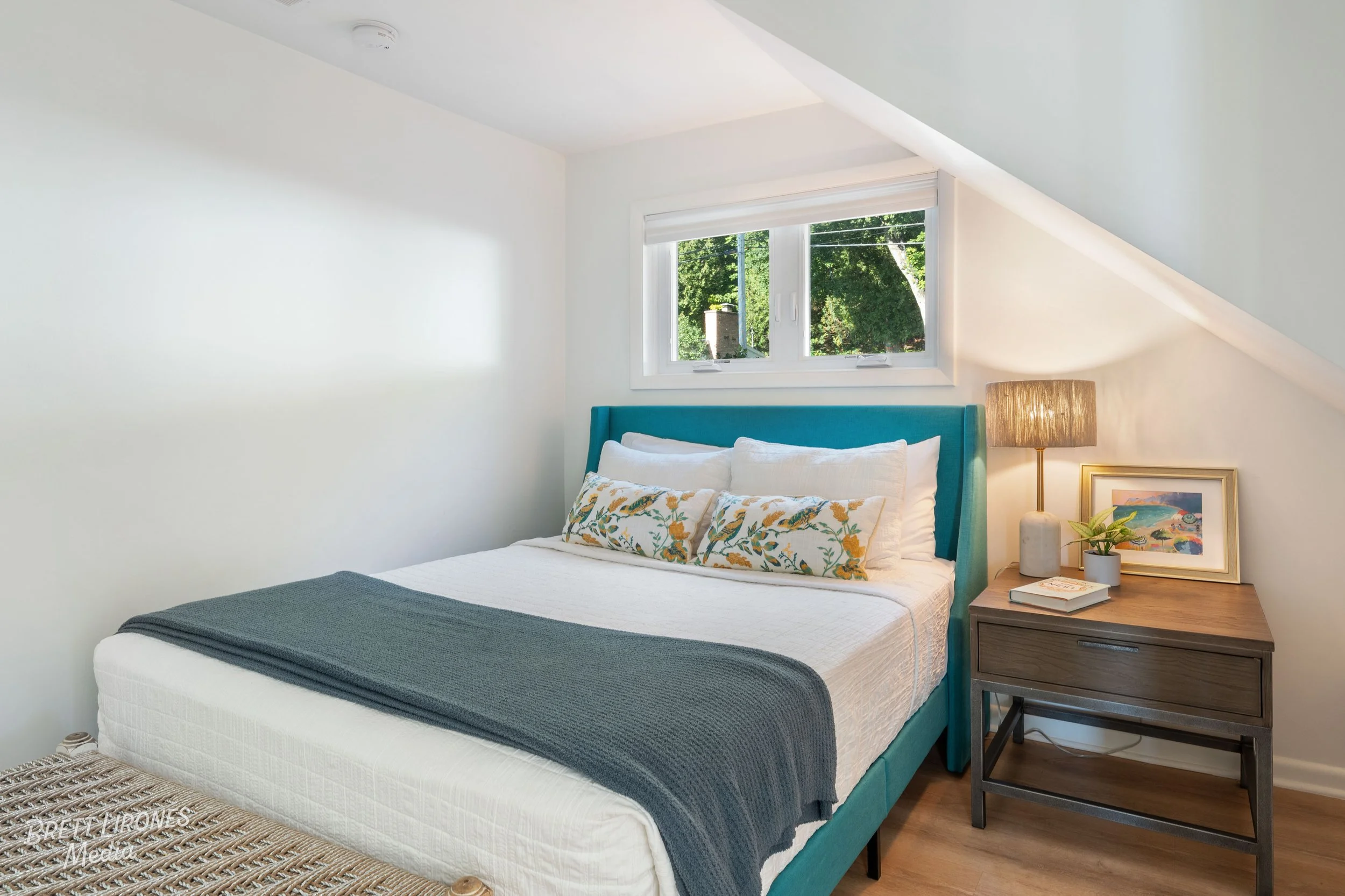 A bedroom with a white wall, a teal upholstered bed with white and floral pillows, a window showing trees outside, a wooden nightstand with a lamp, framed artwork, and a small plant.