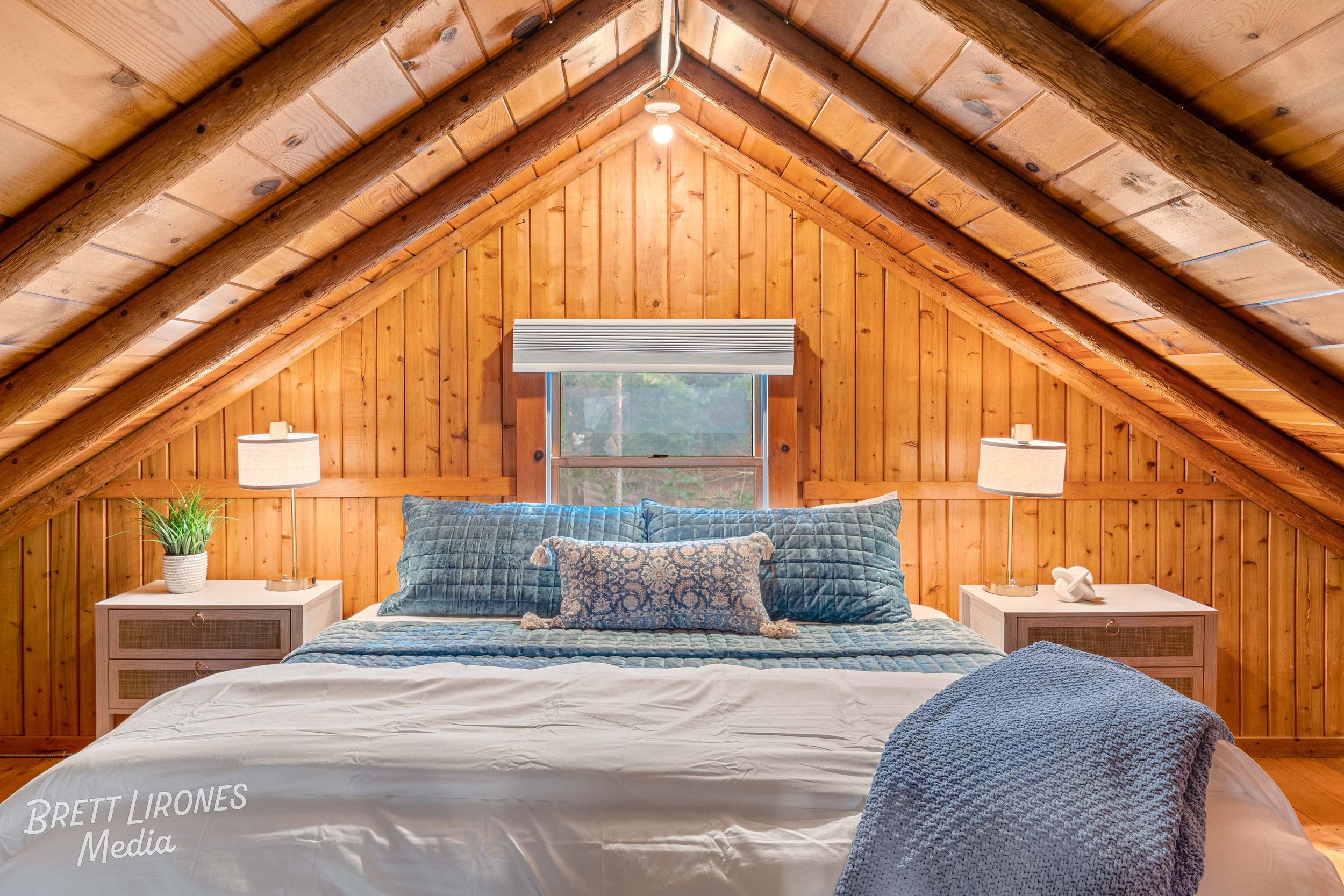 A cozy attic bedroom with wooden paneling, a bed with blue pillows and a patterned decorative pillow, two white nightstands with lamps, a small potted plant, and a window at the back.