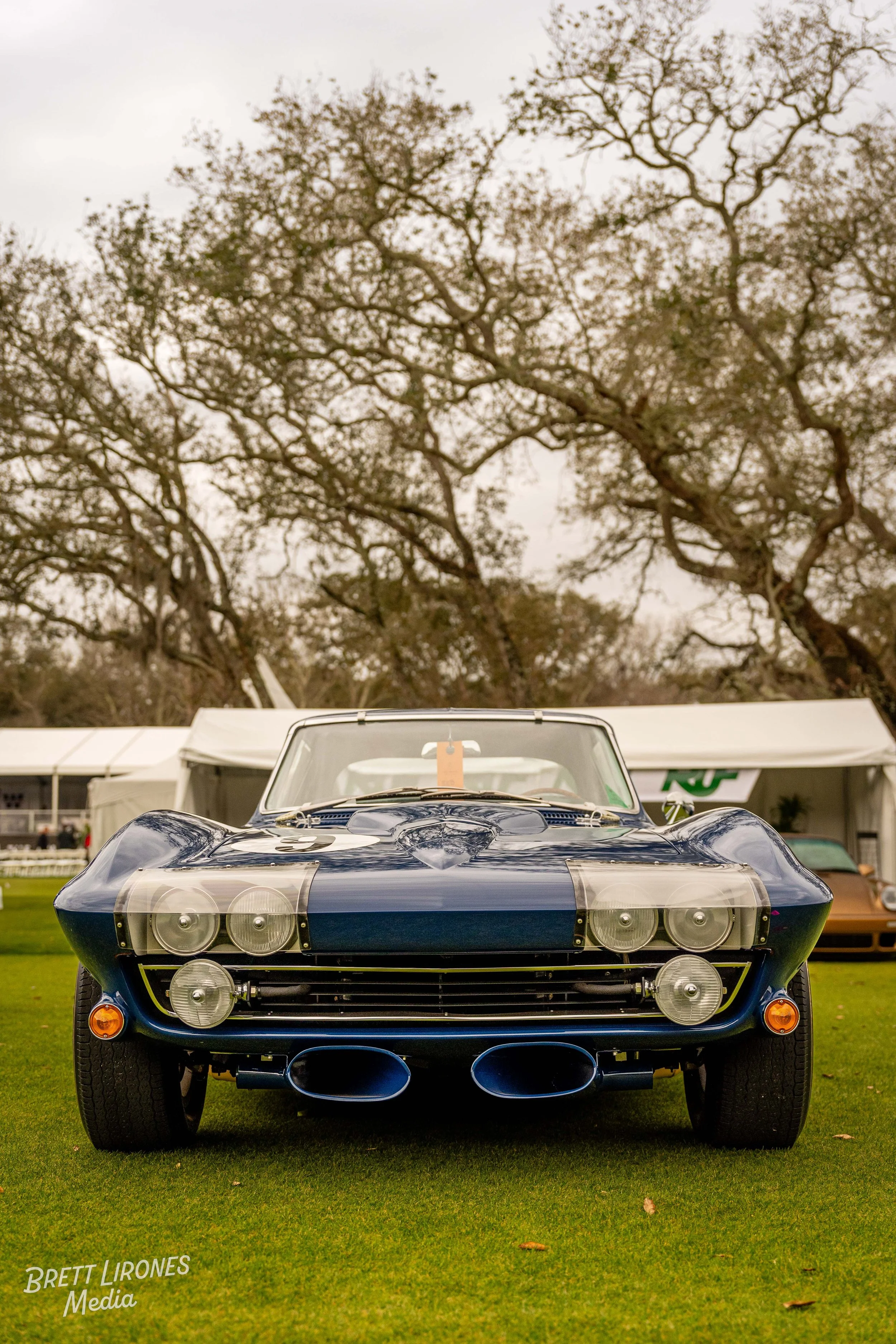 A vintage blue race car displayed outdoors at a car show, with trees and tents in the background.