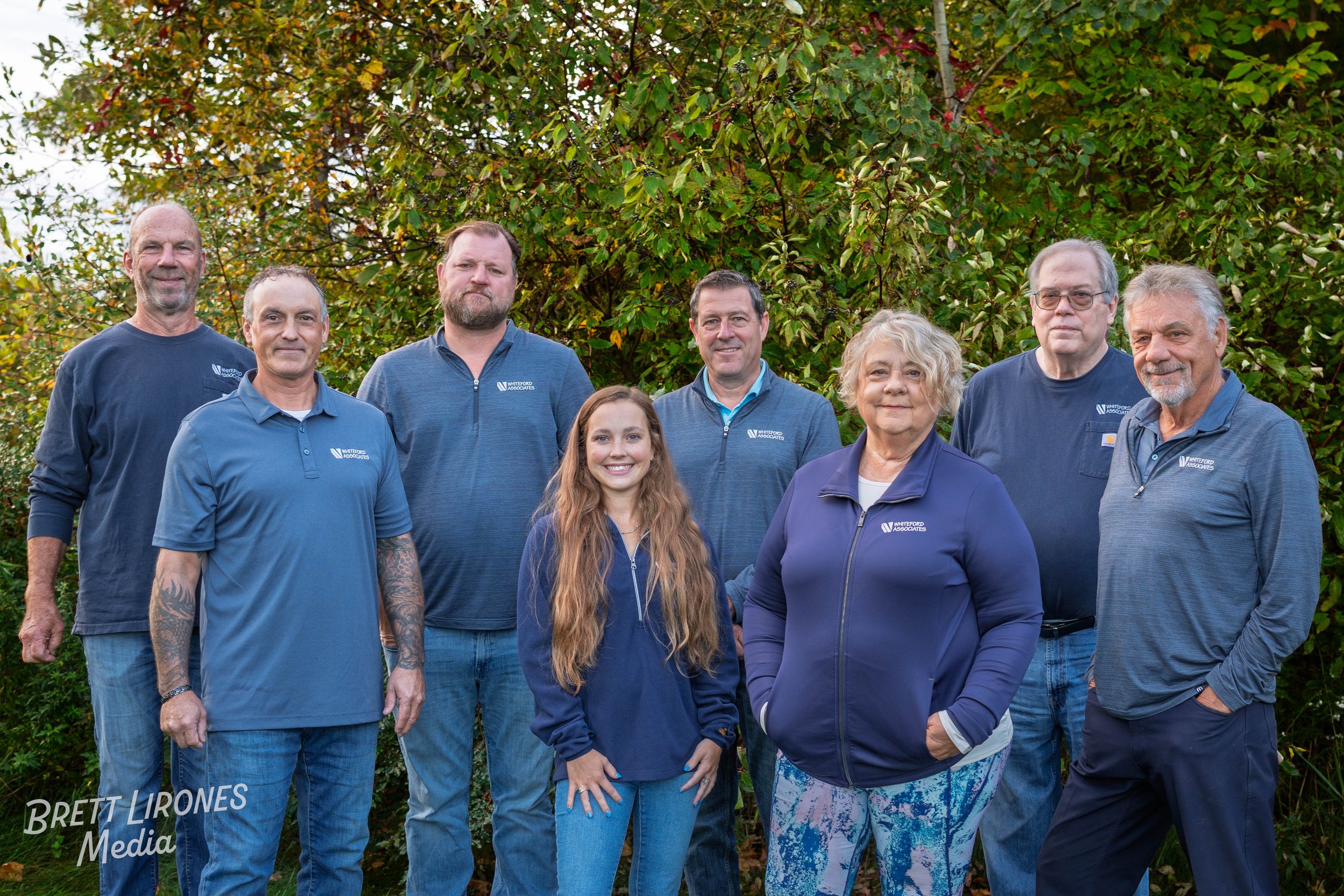 Group of nine people standing outdoors in front of green foliage, dressed in casual and navy blue clothing with company logos, smiling at the camera.