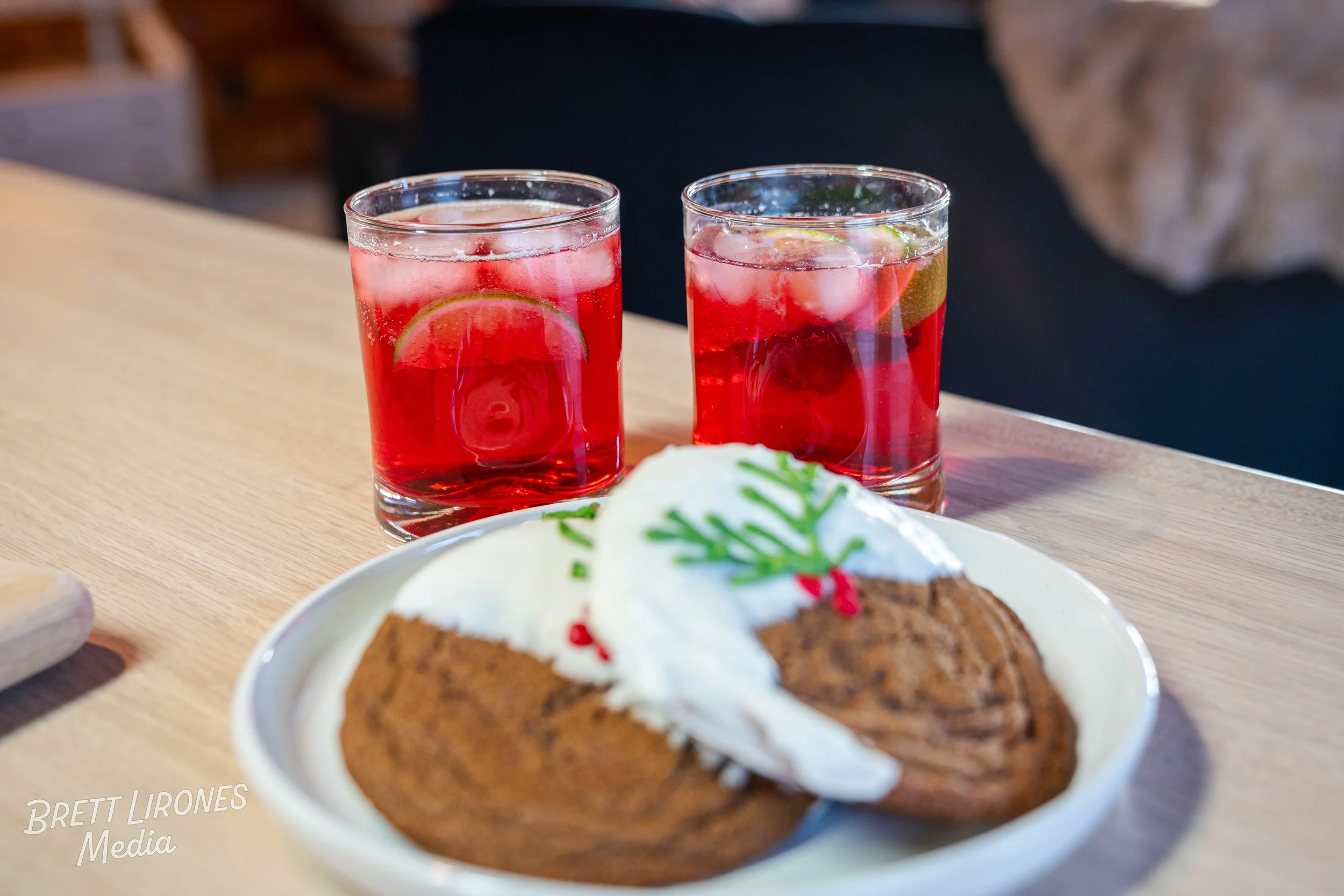 Two glasses of red holiday punch with lime slices on a wooden table, with a plate of cinnamon roll cake topped with whipped cream and festive decorations in the foreground.