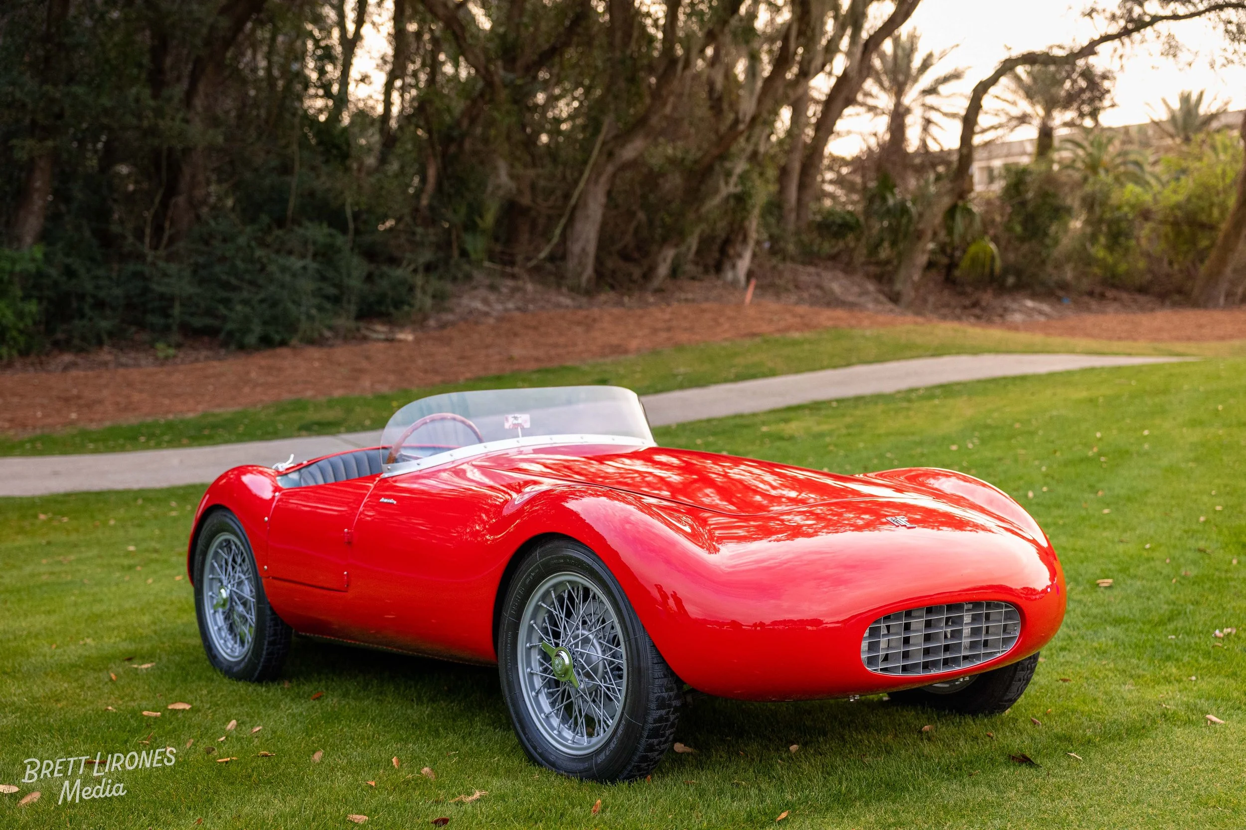 Classic red vintage sports car parked on a grassy area with trees and a walking path in the background.