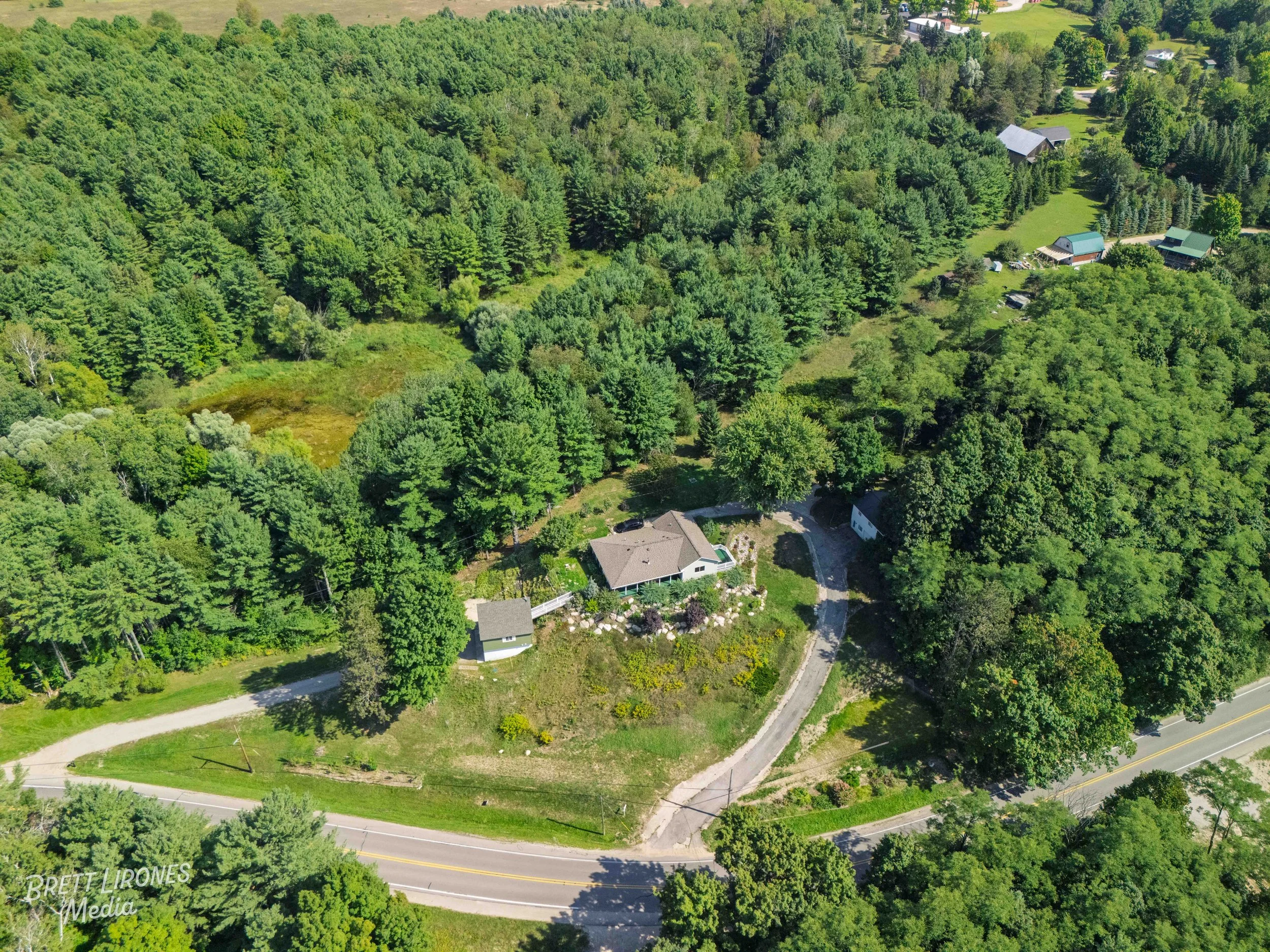 Aerial view of a house surrounded by trees, with a circular driveway, a small outbuilding, and a gravel driveway connecting to a two-lane road. Nearby are other houses and dense forest.