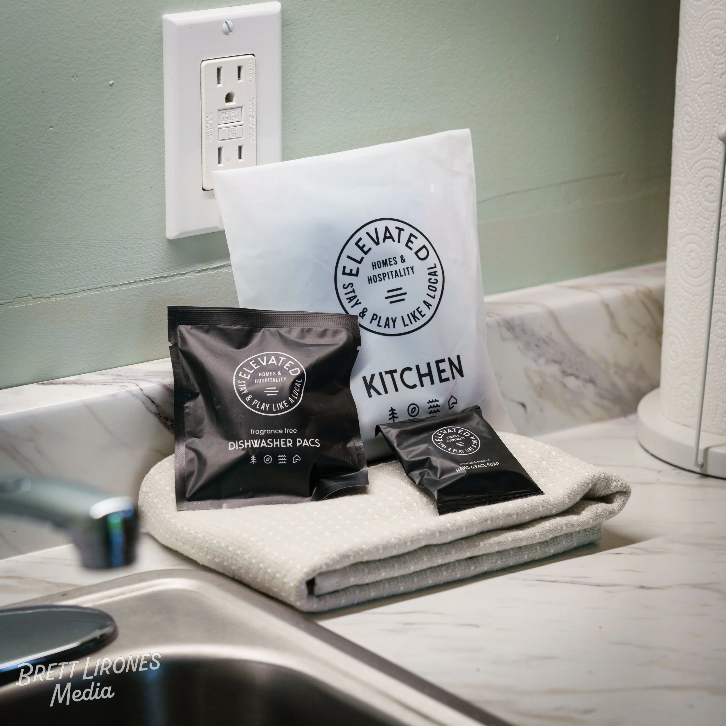 Indoor scene showing a kitchen countertop with coffee bags, above a wall with a double electrical outlet and a paper bag with 'Stay Elevated' printed on it.