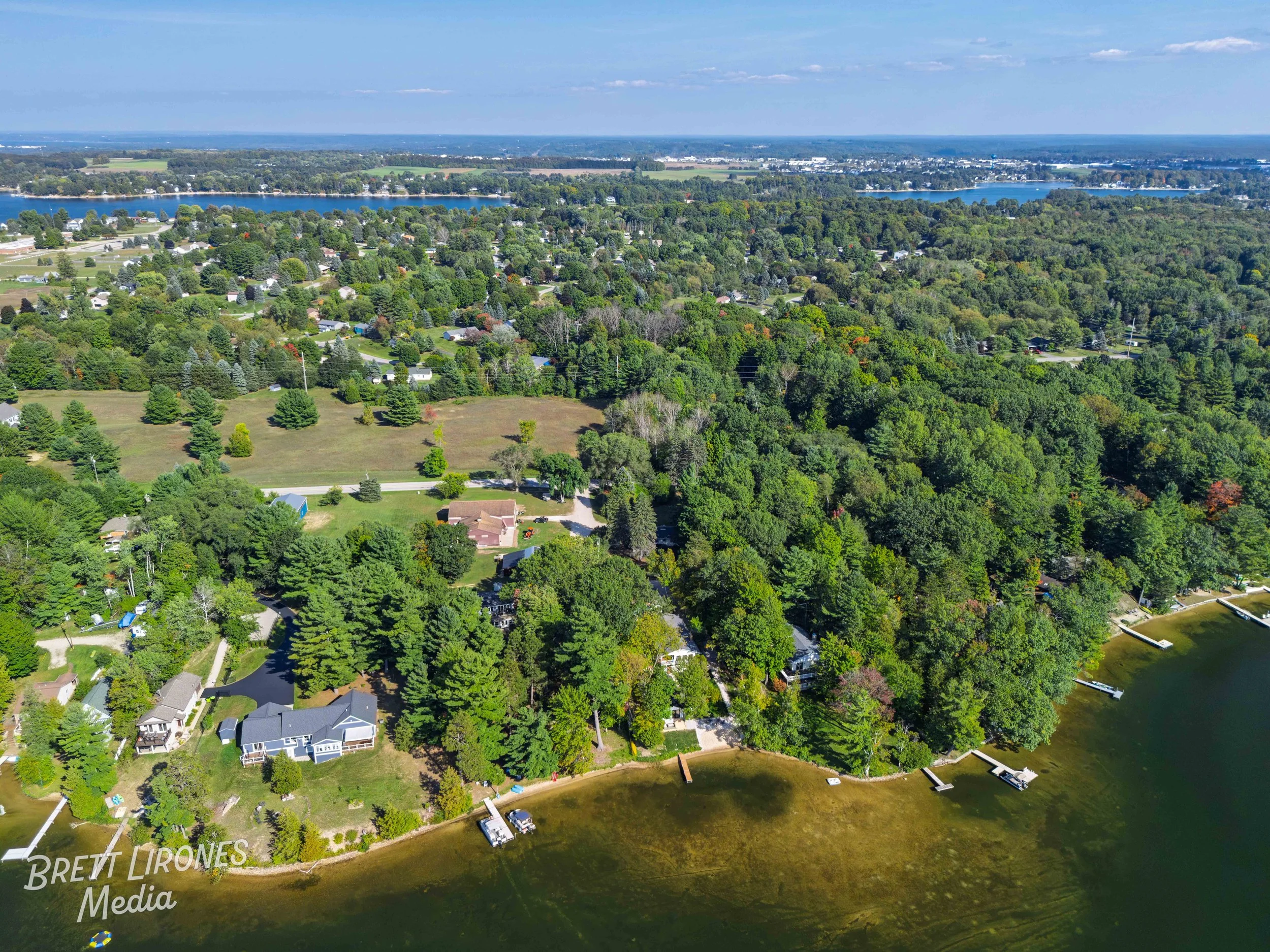 Aerial view of a lush green lakeside neighborhood with houses, trees, and docks along the shoreline. The lake water is calm and the scene is under a partly cloudy sky.