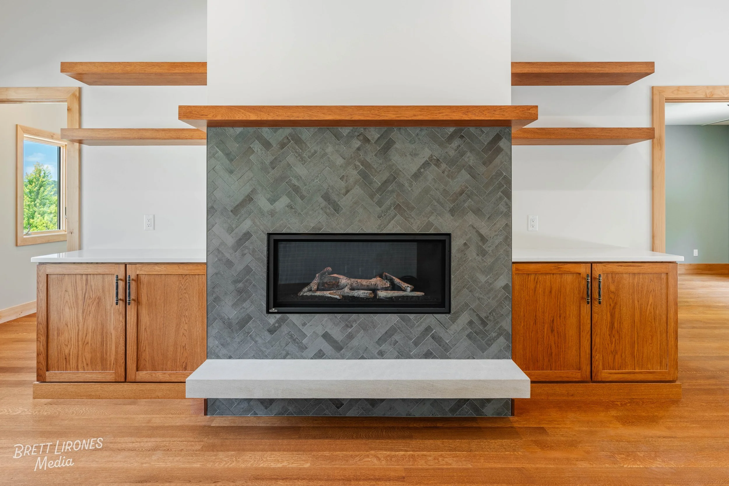 Living room with a modern fireplace with a black frame, surrounded by gray herringbone tile, and wooden shelves and cabinets on either side.