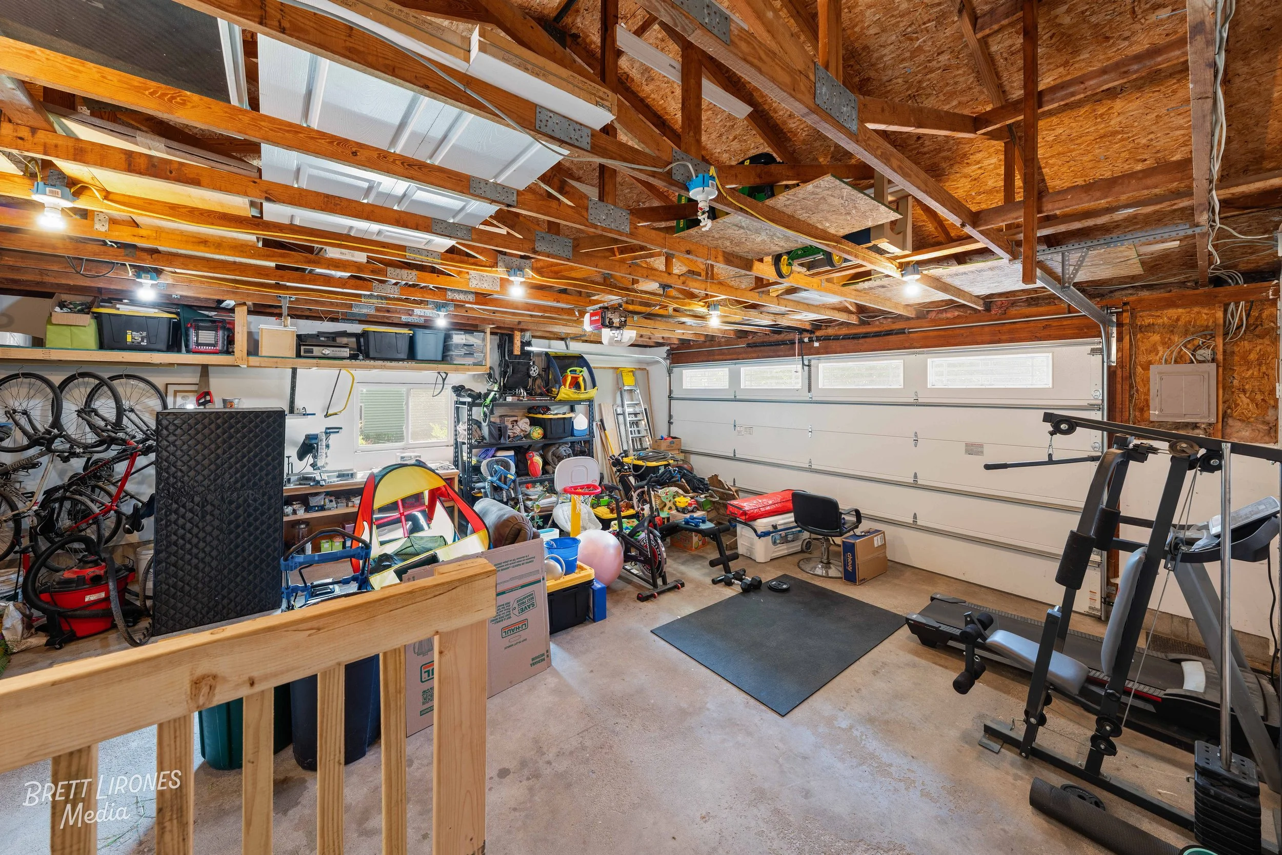 A cluttered garage with a concrete floor, a variety of storage boxes, bikes, workout equipment, and toys, with a partially finished ceiling.