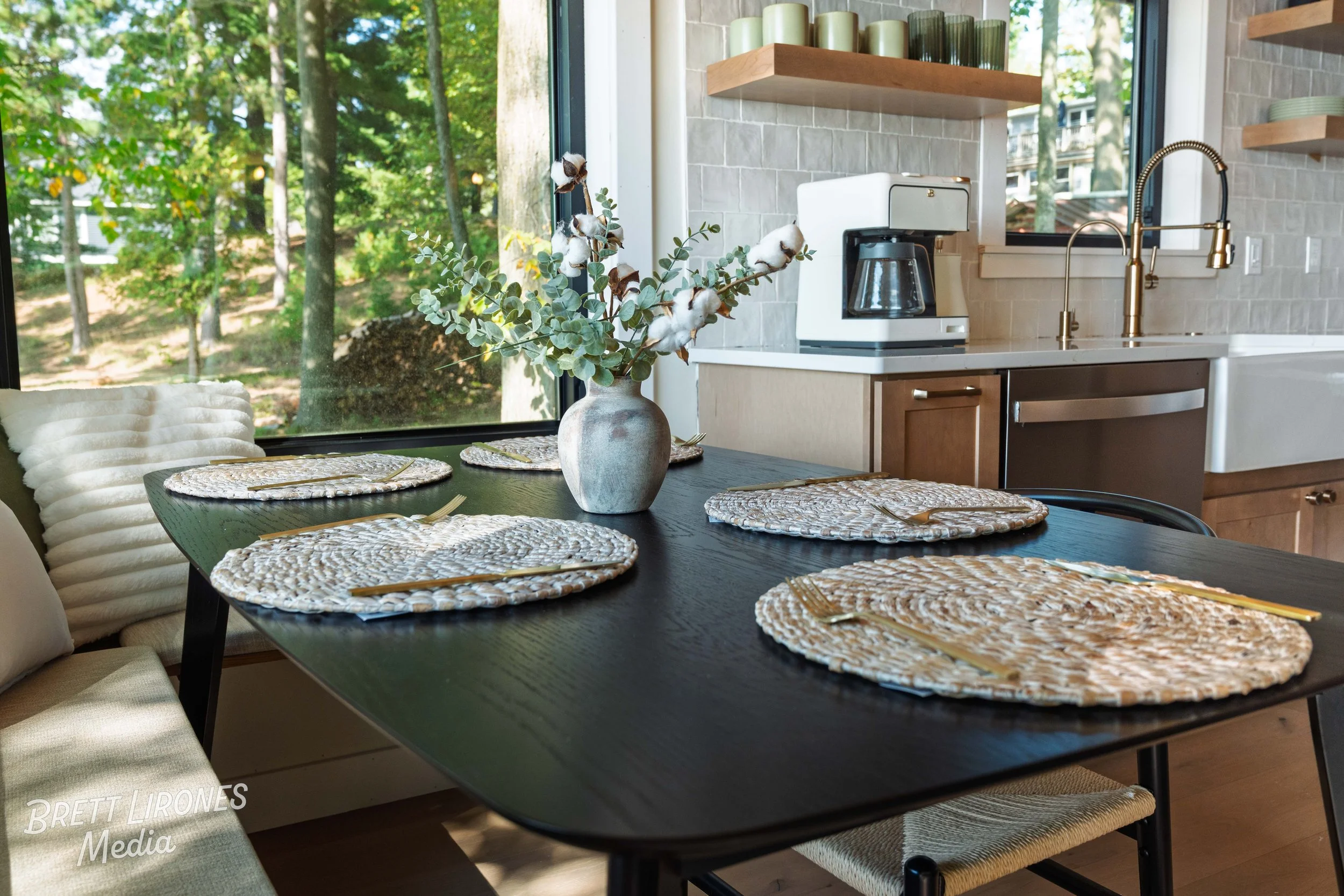 Dining table with woven placemats, a vase with eucalyptus and cotton stems, set in a kitchen with a coffee maker, sink, and window showing trees outside.