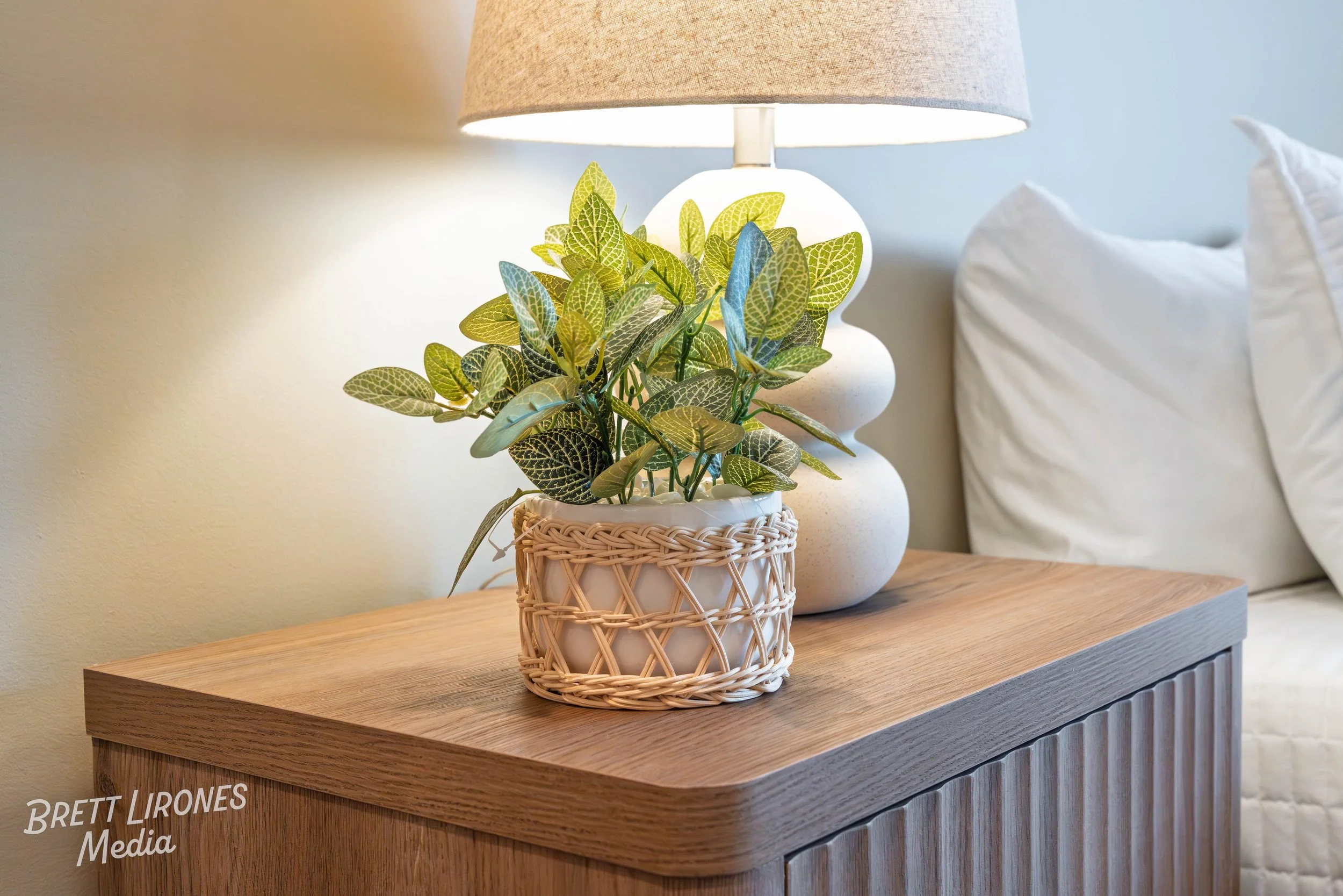 A potted plant with green and yellow variegated leaves on a wooden bedside table, with a white lamp that has a rounded base and beige shade, bedside pillows in the background.