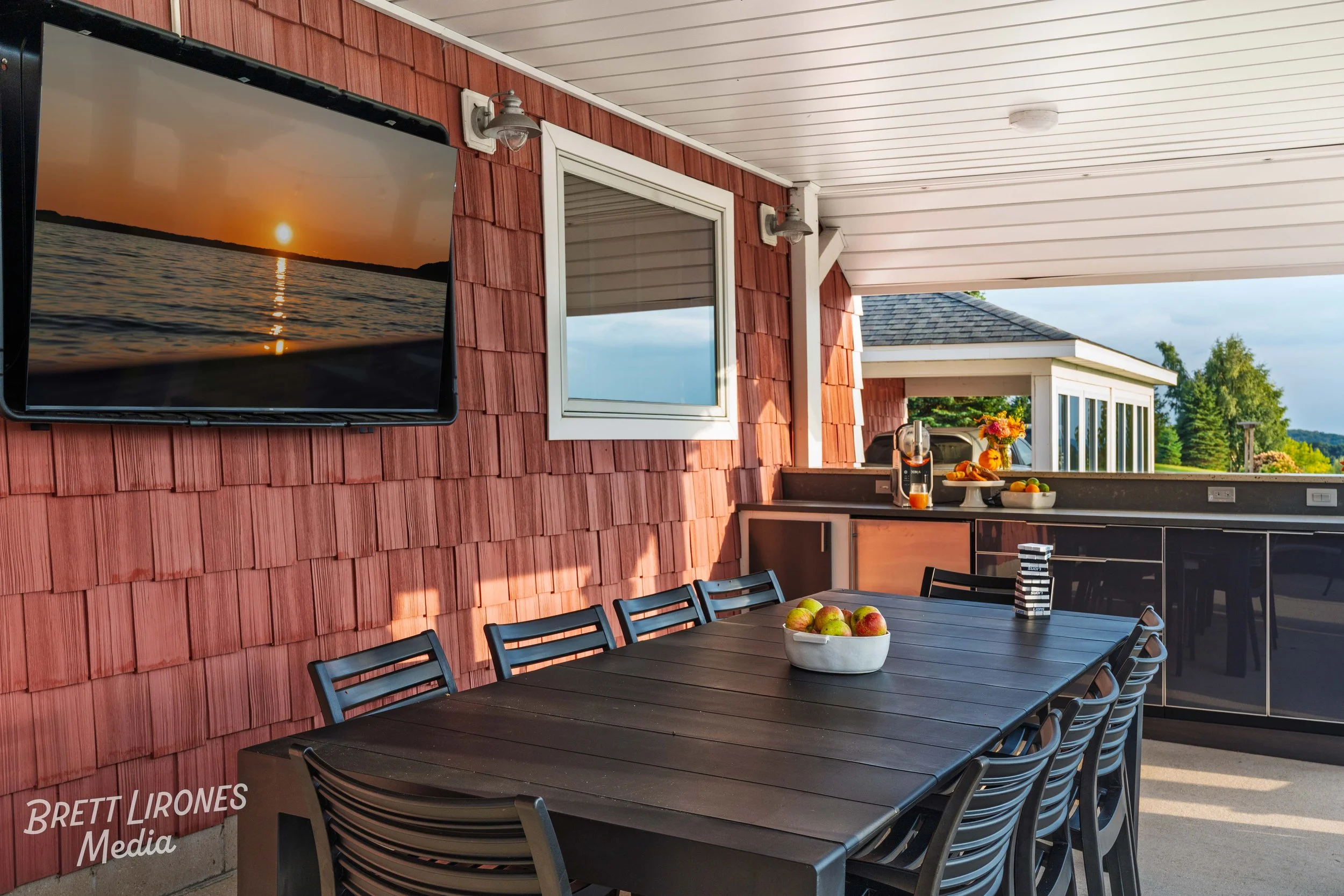 Outdoor patio with a dining table and chairs, a wall-mounted TV showing a sunset over water, a window, and a kitchen counter with a juicer, fruit bowl, and flowers, overlooking a lush green landscape.