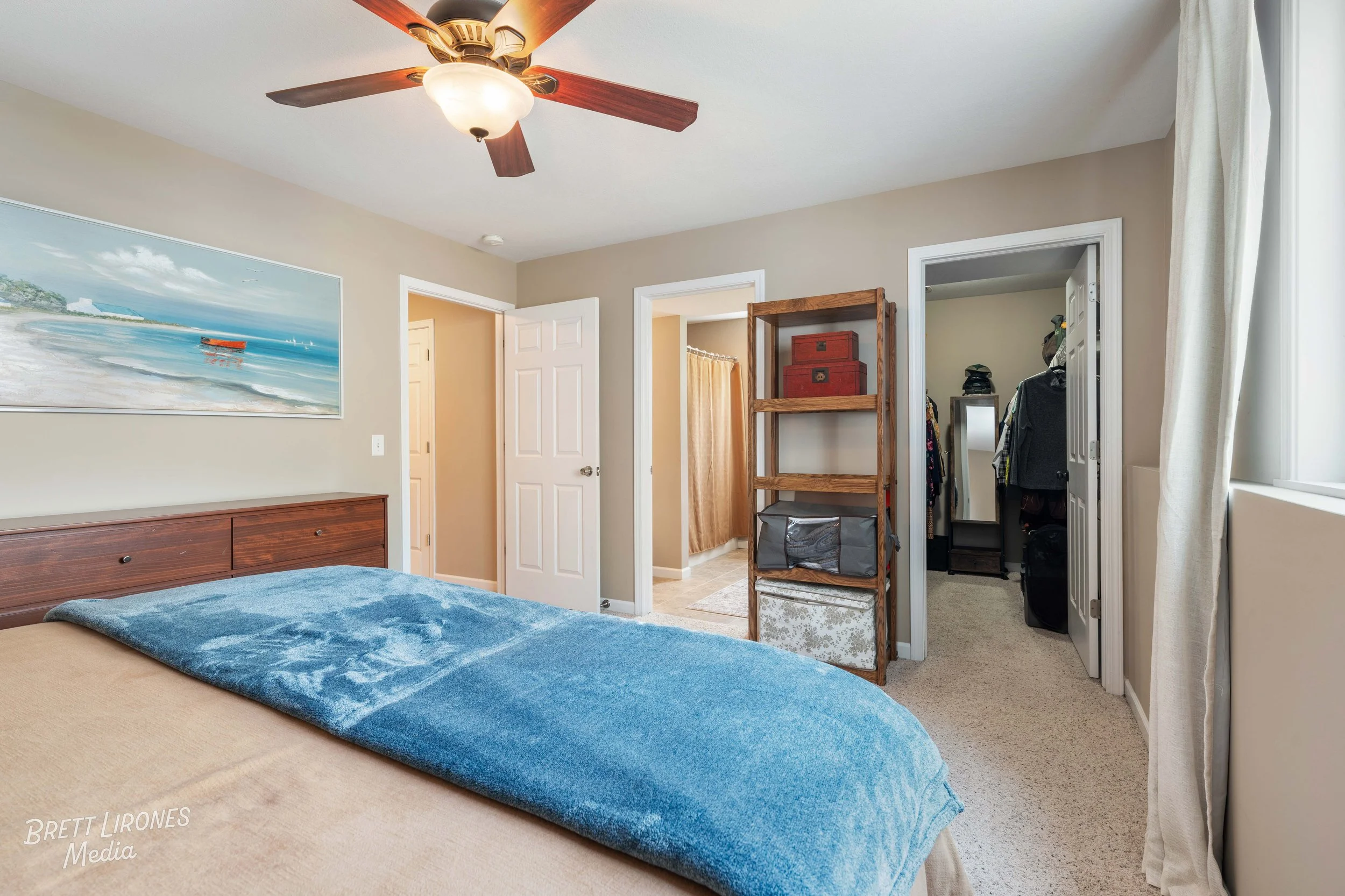 A bedroom with a bed covered by a blue blanket, a wooden headboard with storage drawers, a ceiling fan with lights, a painting of a beach scene on the wall, and an open closet with clothes and a mirror.