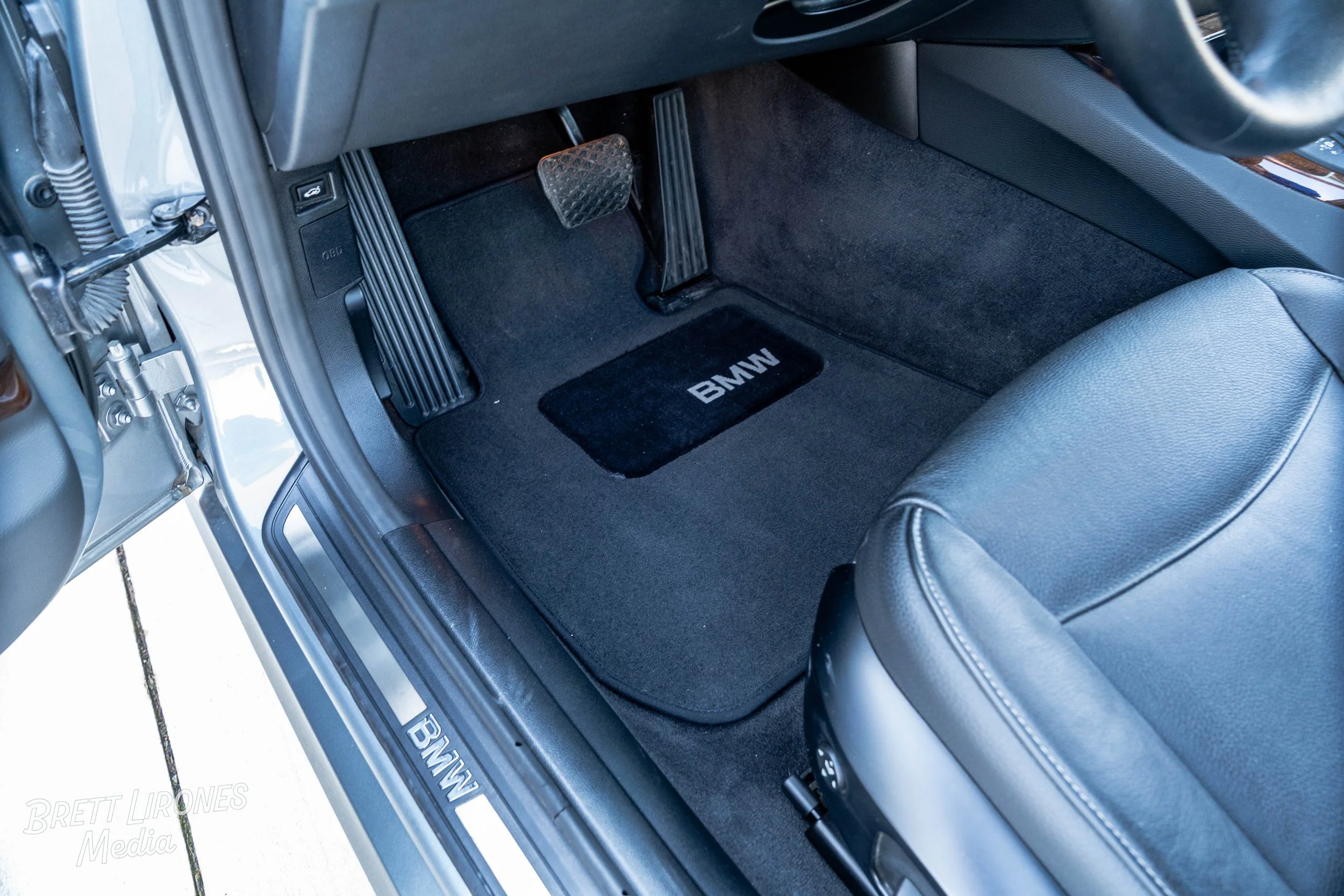 The interior of a BMW car shows the driver's side floor area with a black carpeted mat with the BMW logo, an accelerator pedal, and a brake pedal. The seat is black leather, and part of the door sill with the BMW logo is visible.