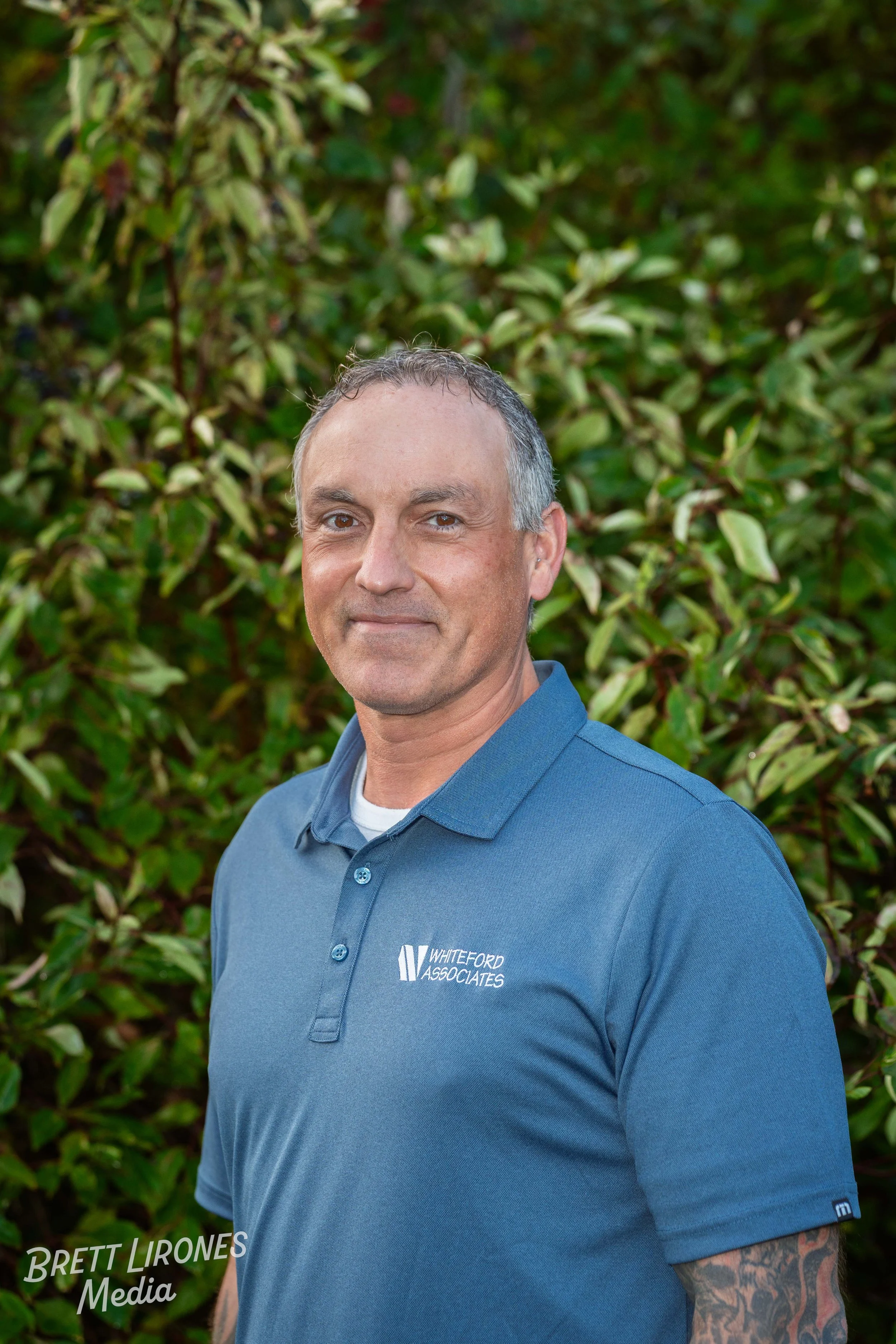 A man in a blue polo shirt standing outdoors in front of green bushes.
