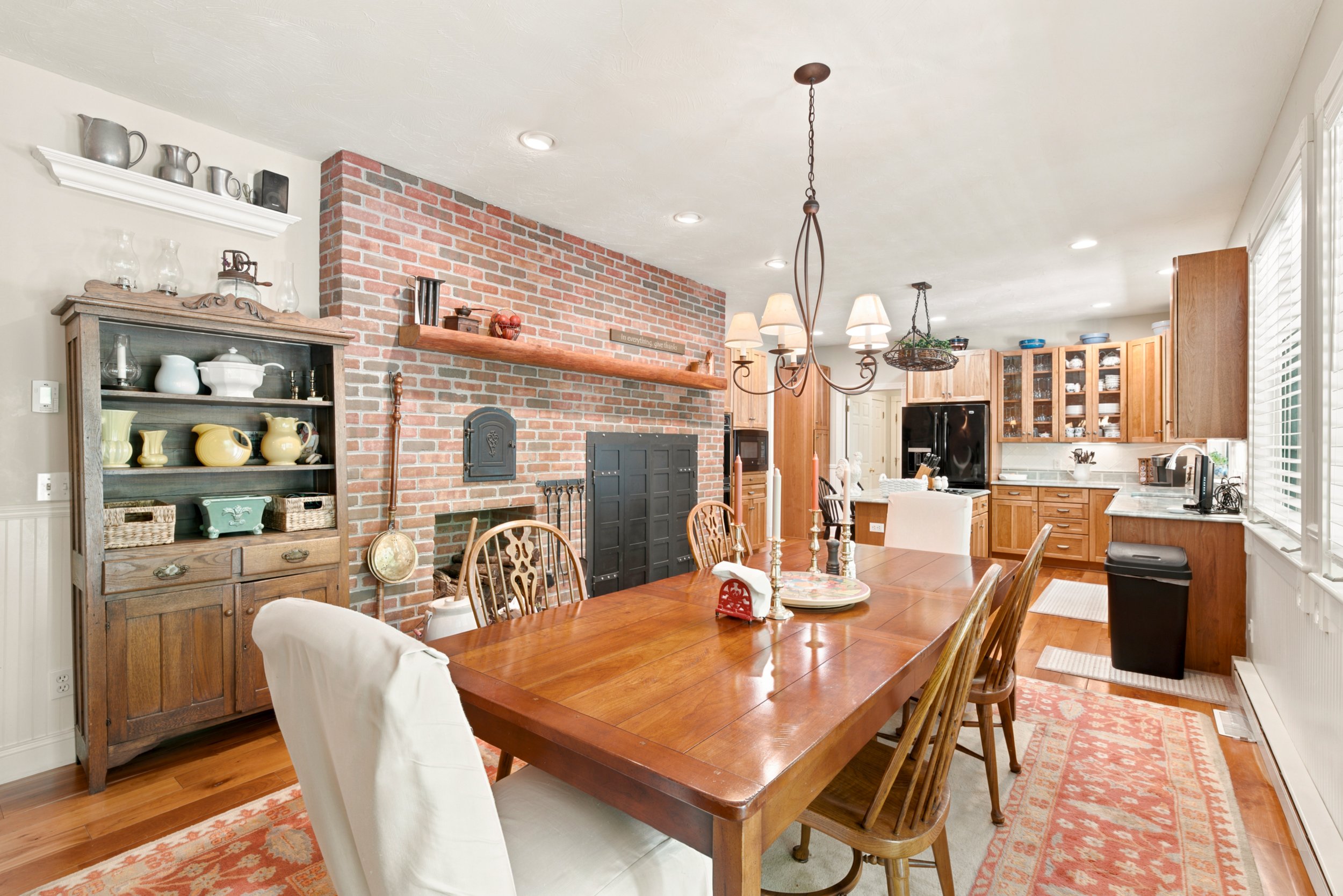 A dining area with a wooden table and chairs, a painted brick fireplace, and kitchen cabinets. There is a shelf with pitchers and glassware, a chandelier above the table, and large windows with white blinds.
