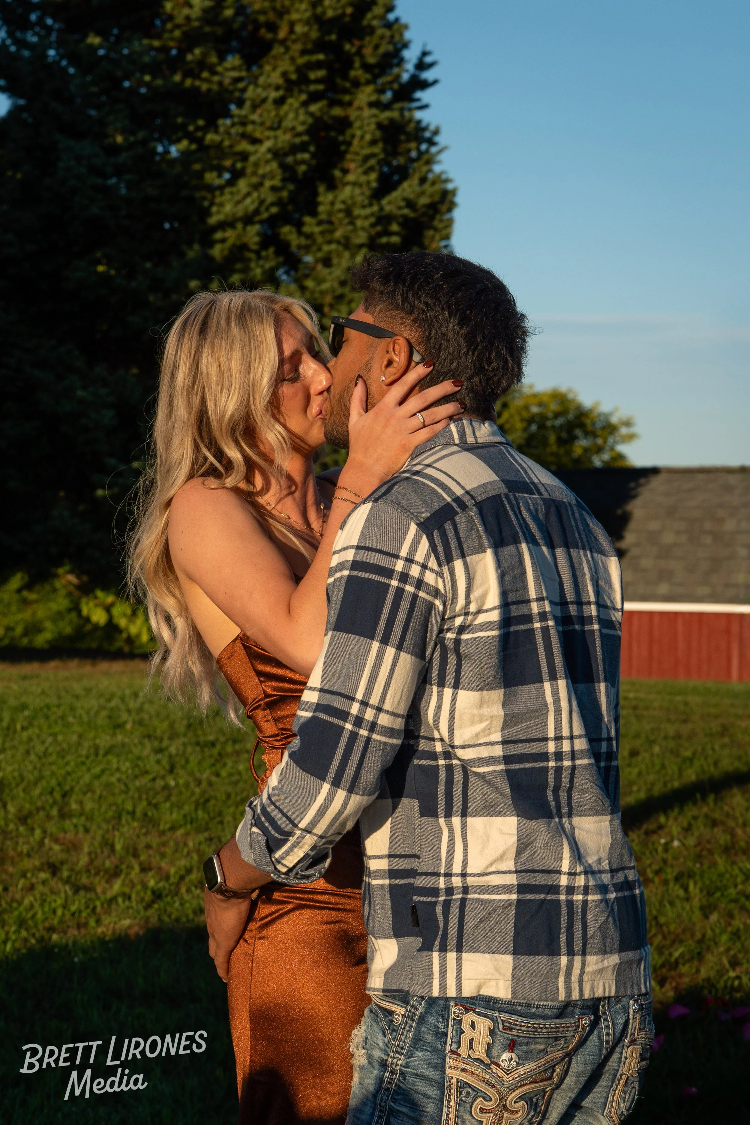 A couple kissing outdoors during sunset, with the woman wearing a copper-colored dress and the man wearing a plaid shirt and jeans, with trees and a building in the background.