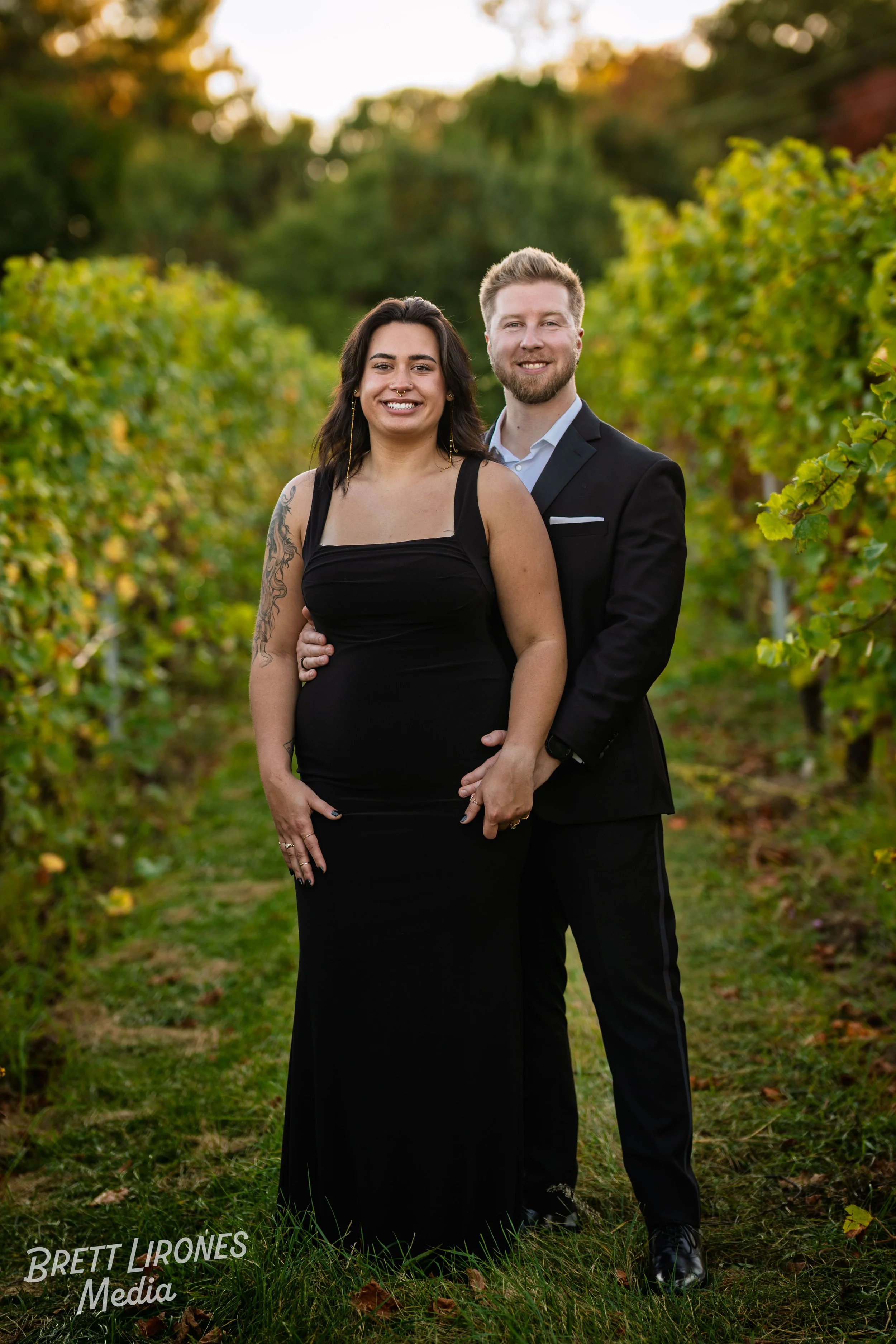 A smiling couple standing hand in hand in a vineyard during sunset, with the woman wearing a black dress and the man in a suit.