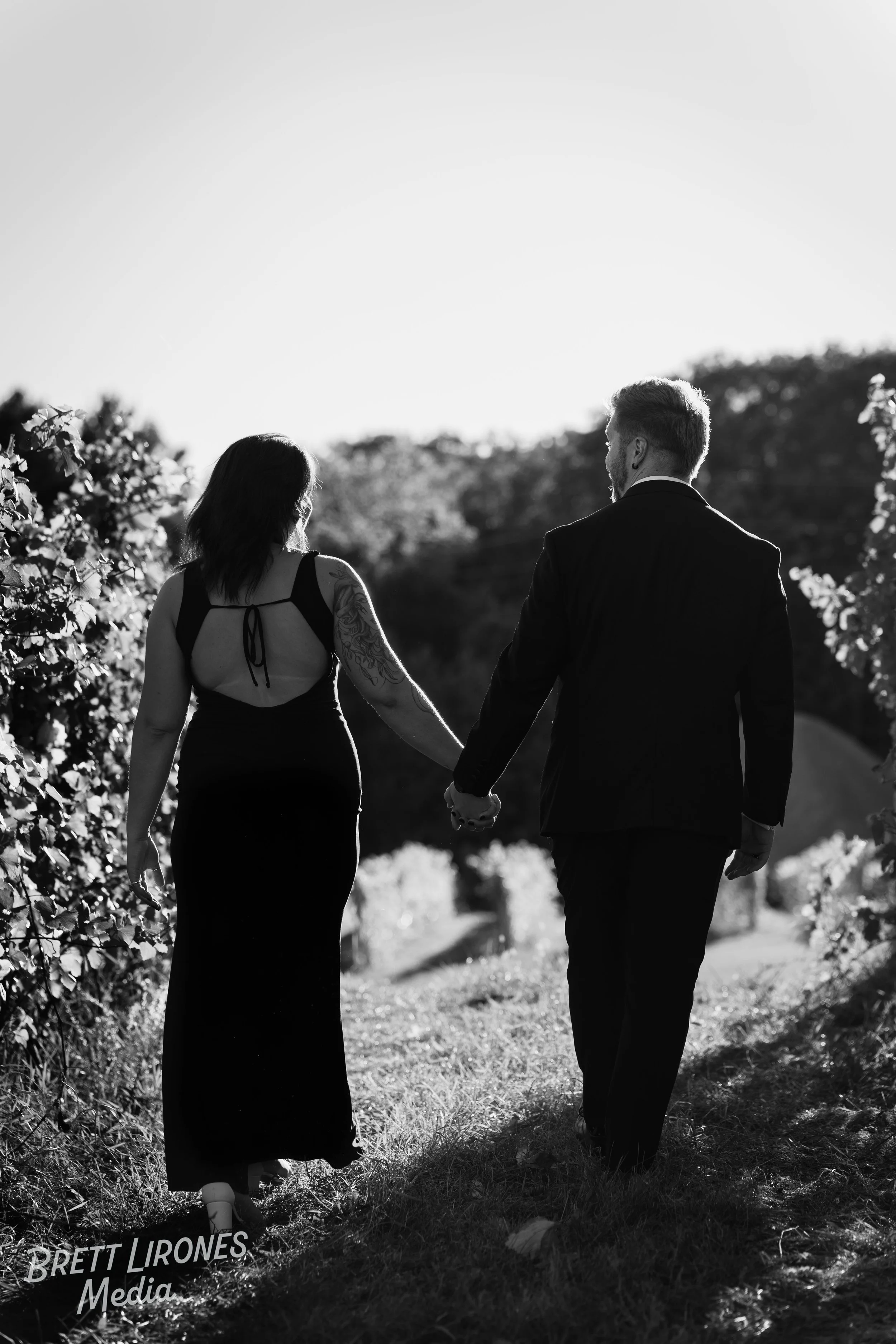 A black and white photo of a couple walking hand in hand through a natural setting, possibly a garden or park, with trees and bushes around them during the daytime.
