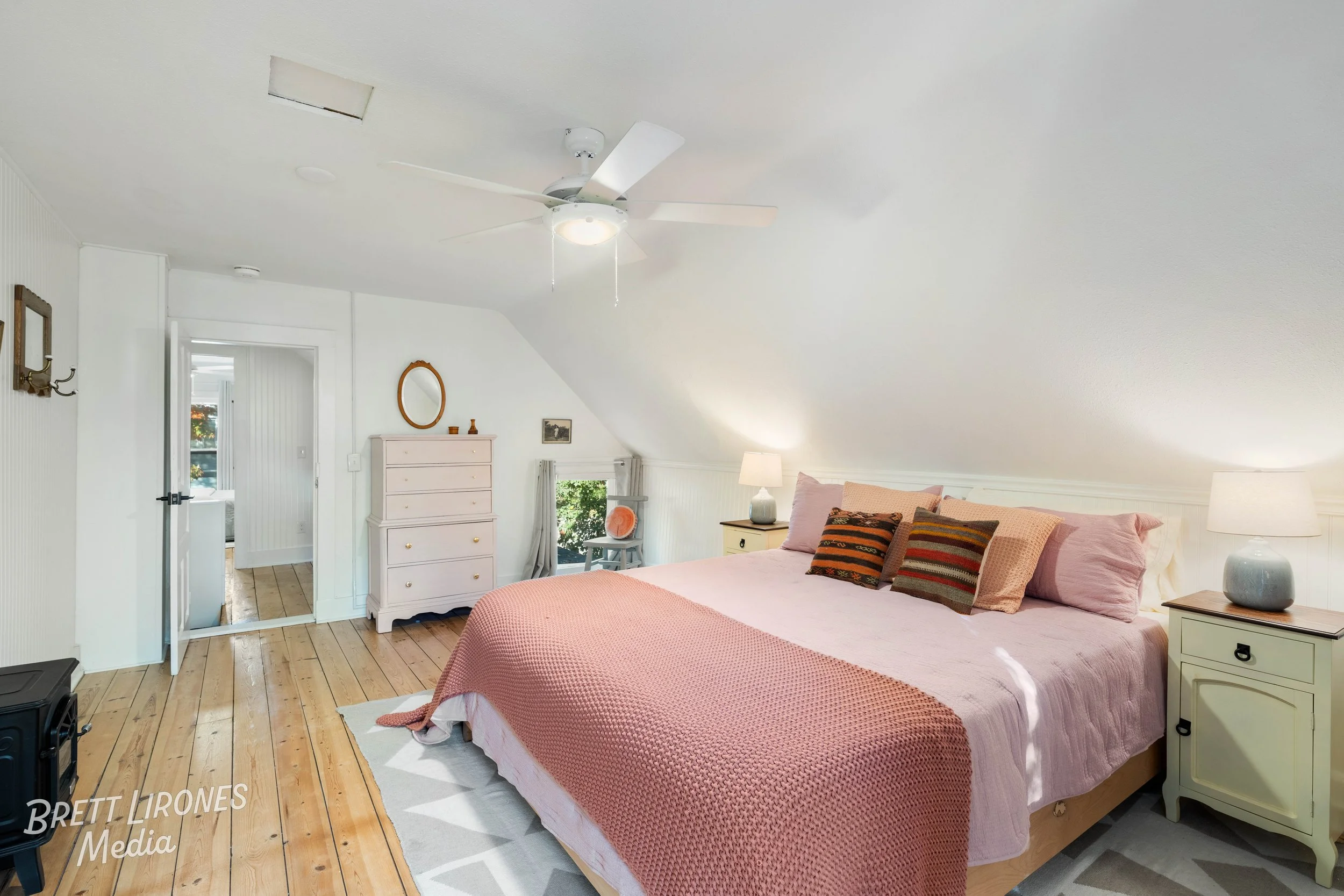 A cozy bedroom with a sloped ceiling, featuring a large bed with pink bedding and decorative pillows, two nightstands with lamps, a small dresser, a ceiling fan, and a window with curtains.