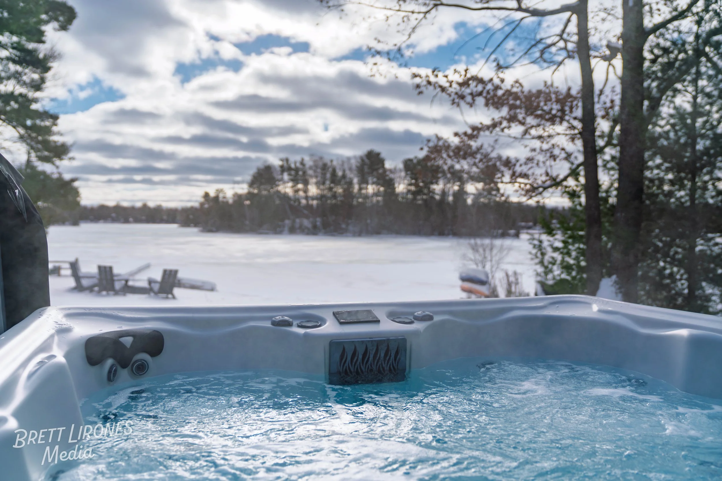 A hot tub filled with water outdoors near a frozen lake, with trees and a cloudy sky in the background.