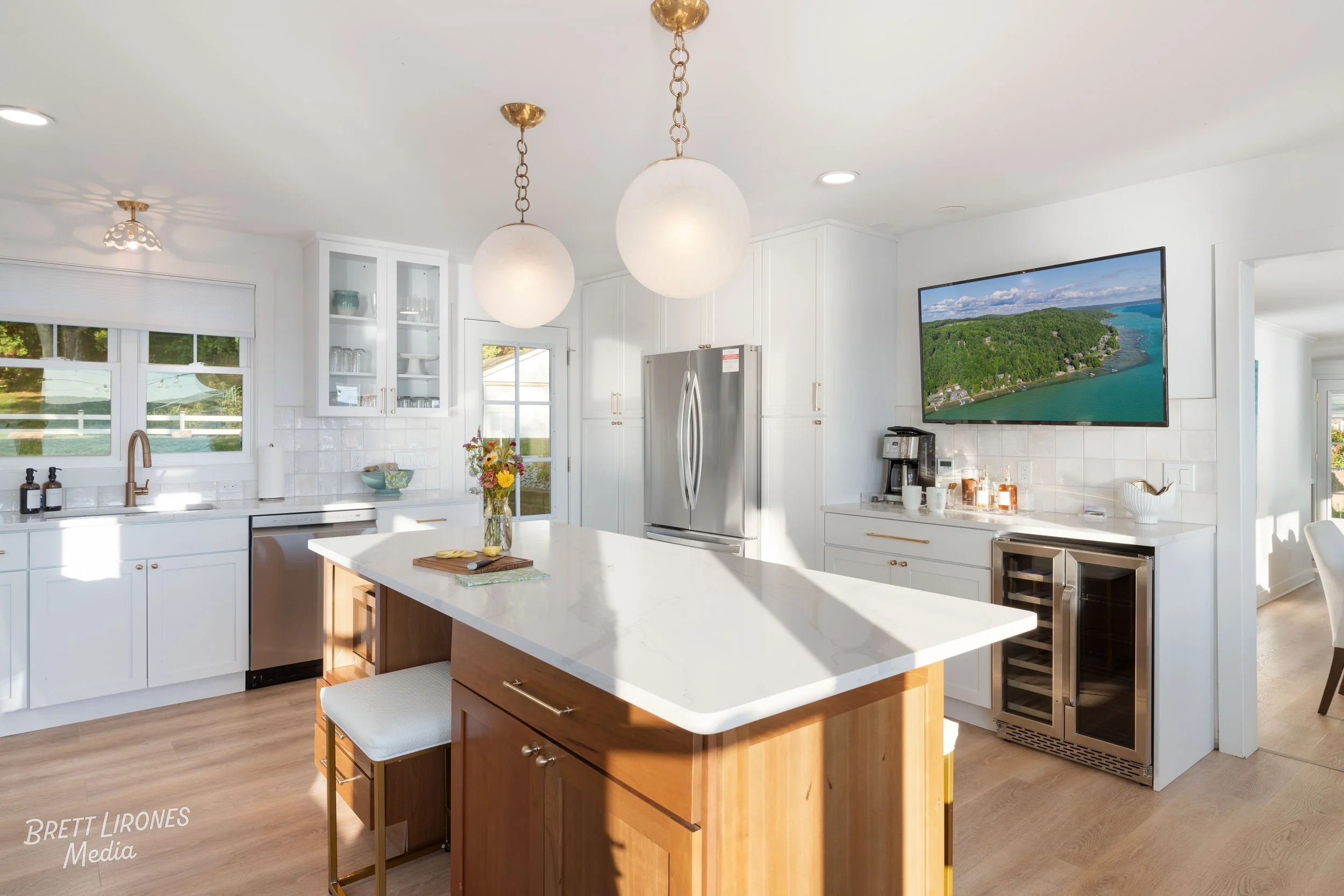 Modern kitchen with white cabinets, a marble countertop island, stainless steel appliances, pendant lights, a wall-mounted TV showing a landscape, and wooden flooring.