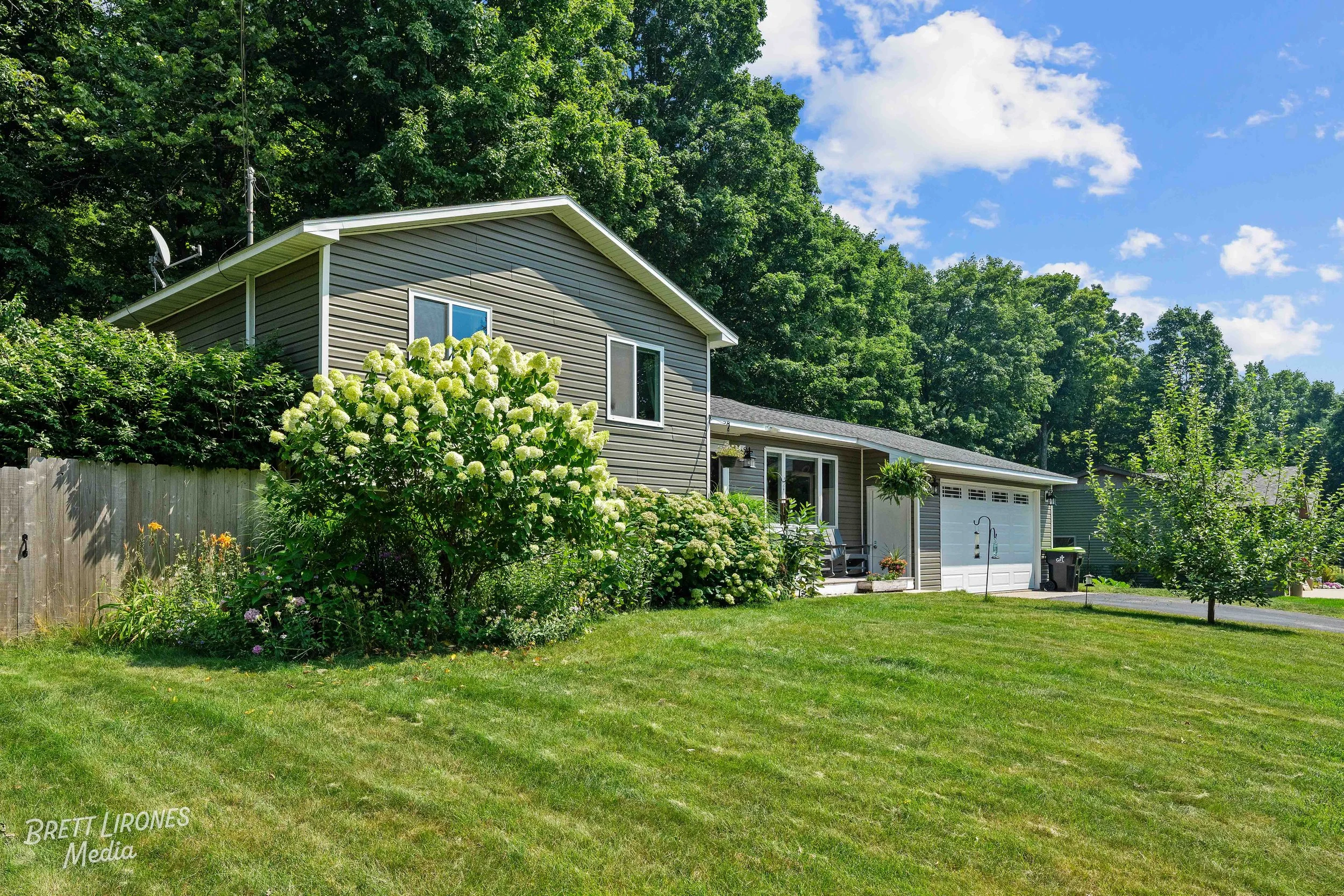 A suburban house with grey siding, a white garage door, and a small front porch decorated with plants. The yard features a well-maintained green lawn, large white-flowered bushes, and a small tree, with tall trees and a blue sky with clouds in the ba