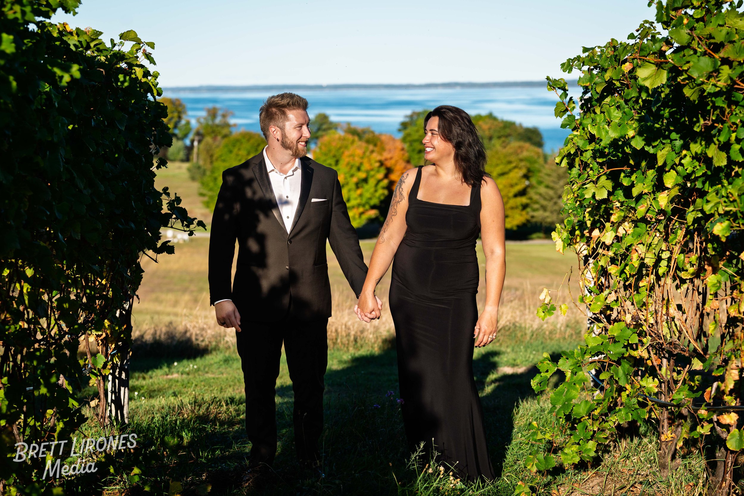 A couple in formal attire walking hand in hand outdoors, surrounded by greenery with a body of water and trees in the background.