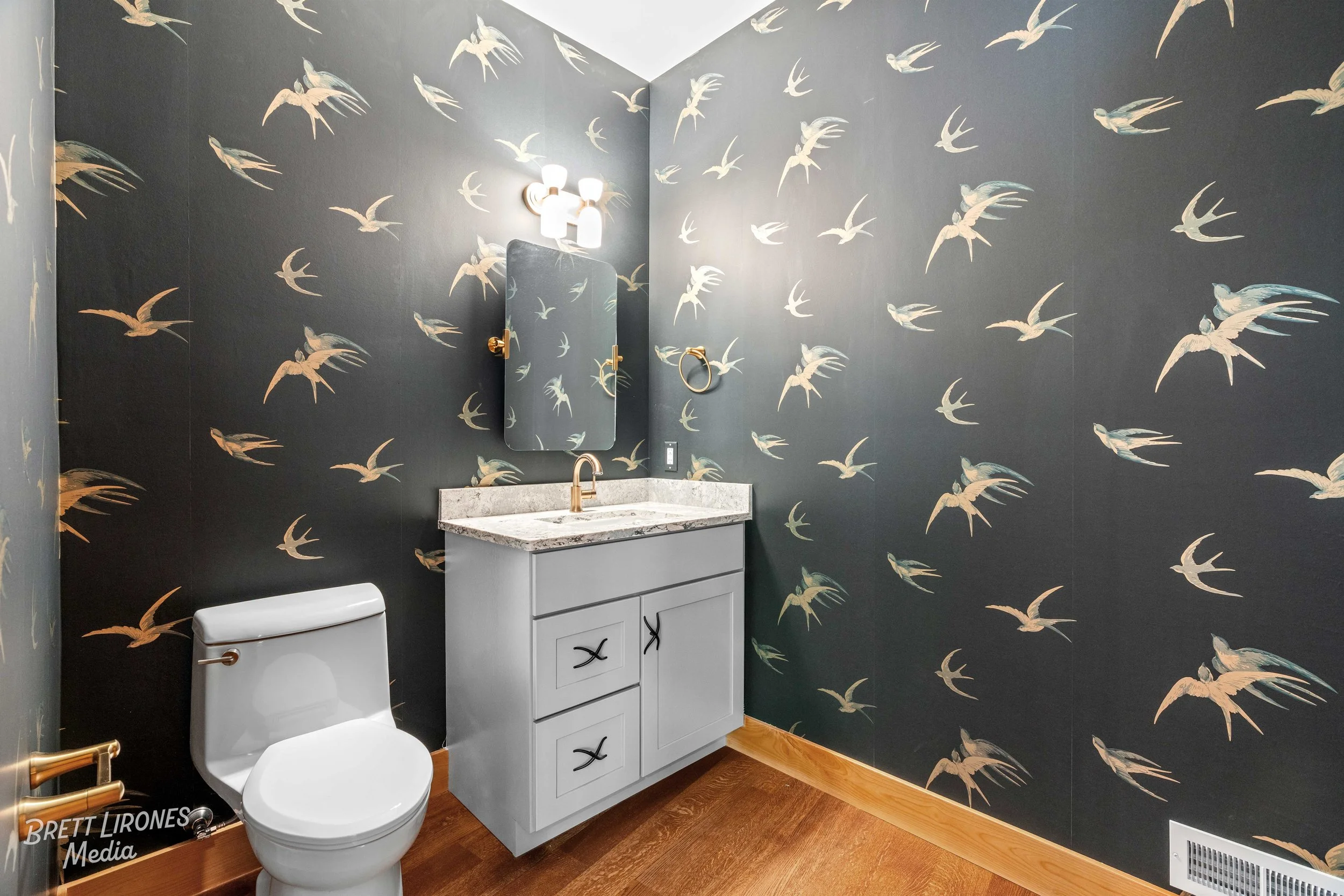 A bathroom with dark wallpaper featuring bird illustrations, a white toilet, a white vanity with a marble countertop, gold fixtures, a mirror, and a wall-mounted light fixture.